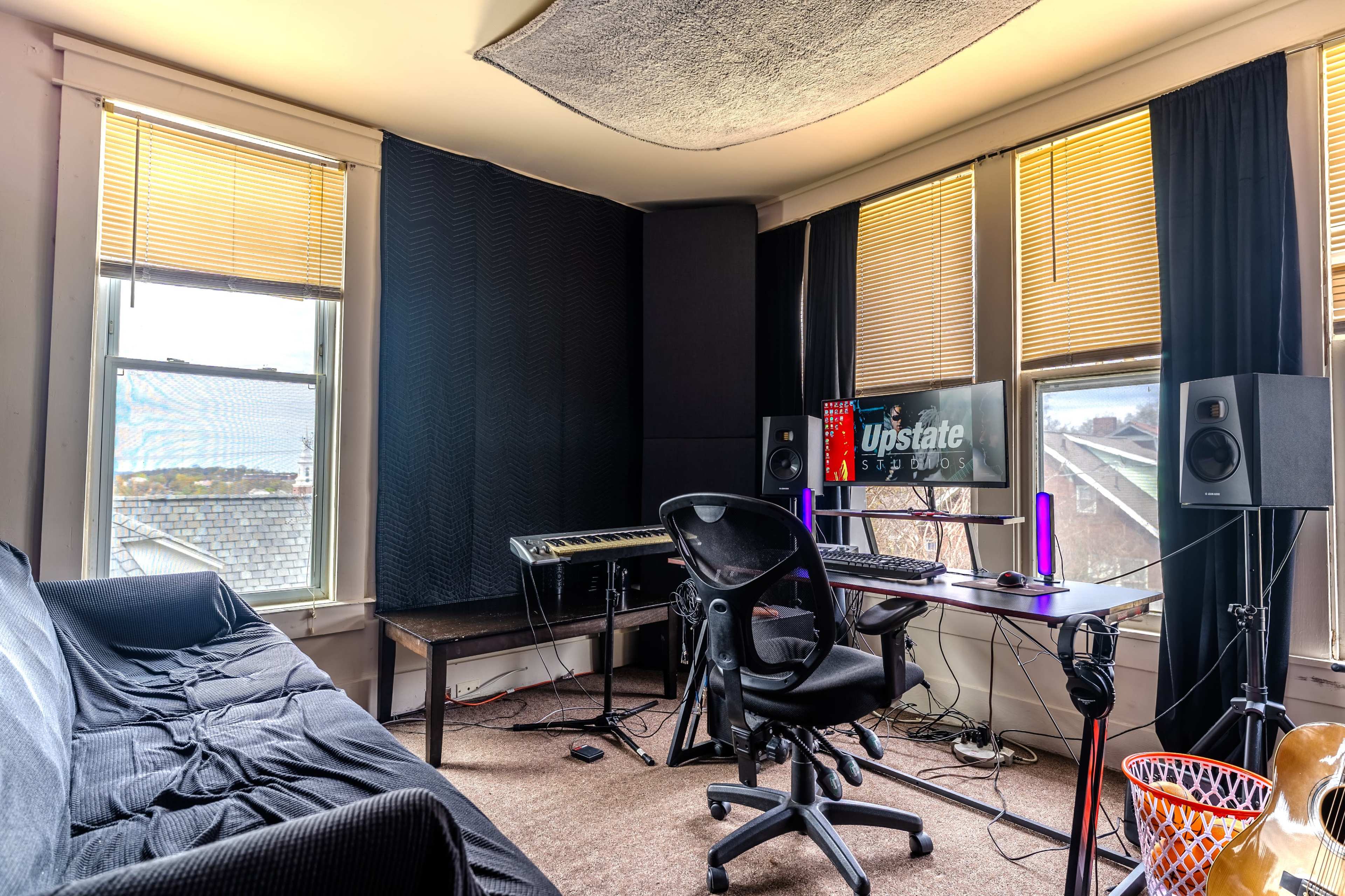 A studio room features a desk with a computer and speakers, a keyboard, a black couch, and large windows with yellow blinds.