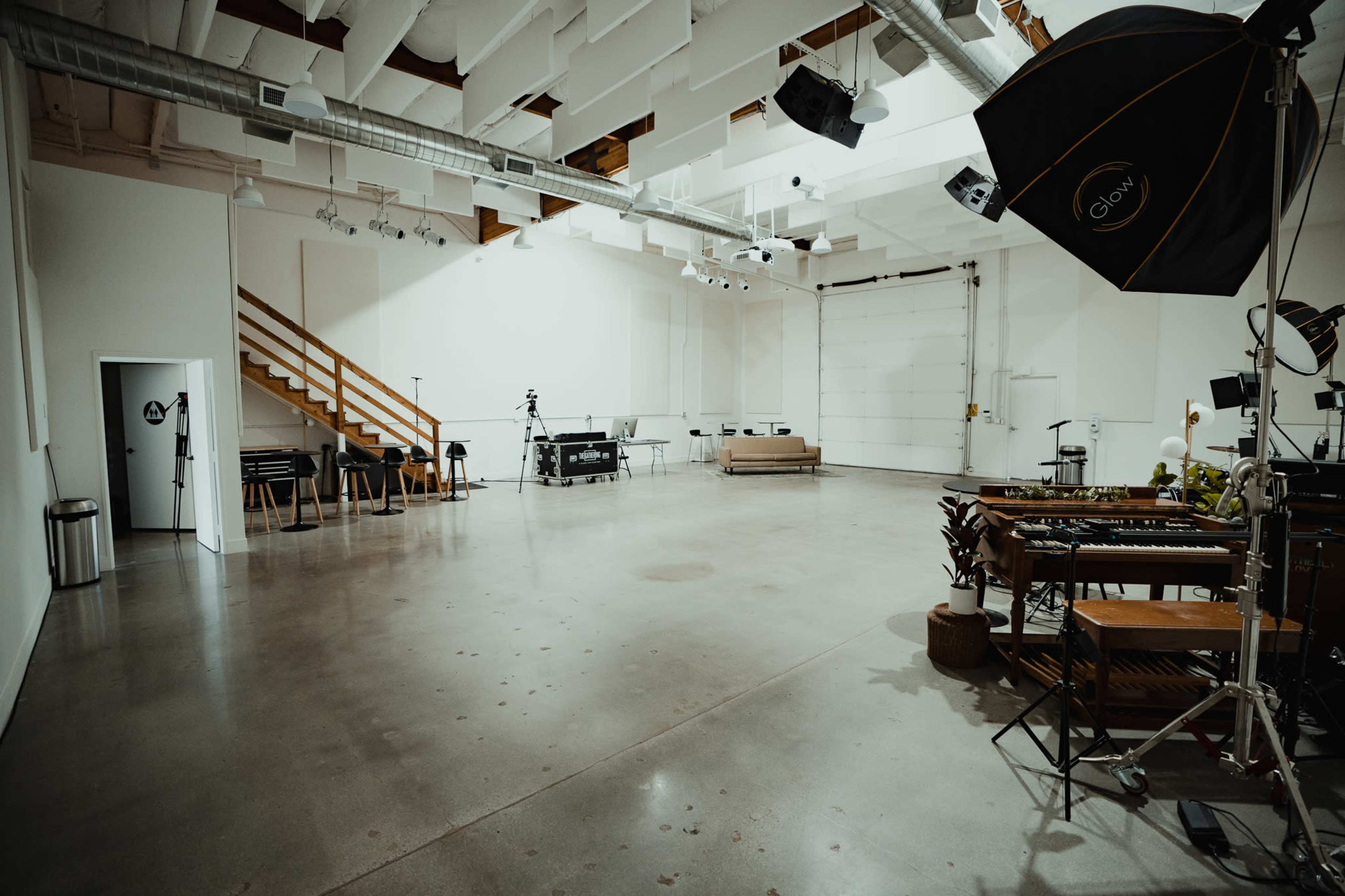 A spacious, well-lit studio features concrete floors, a staircase, and several photography and video equipment setups.