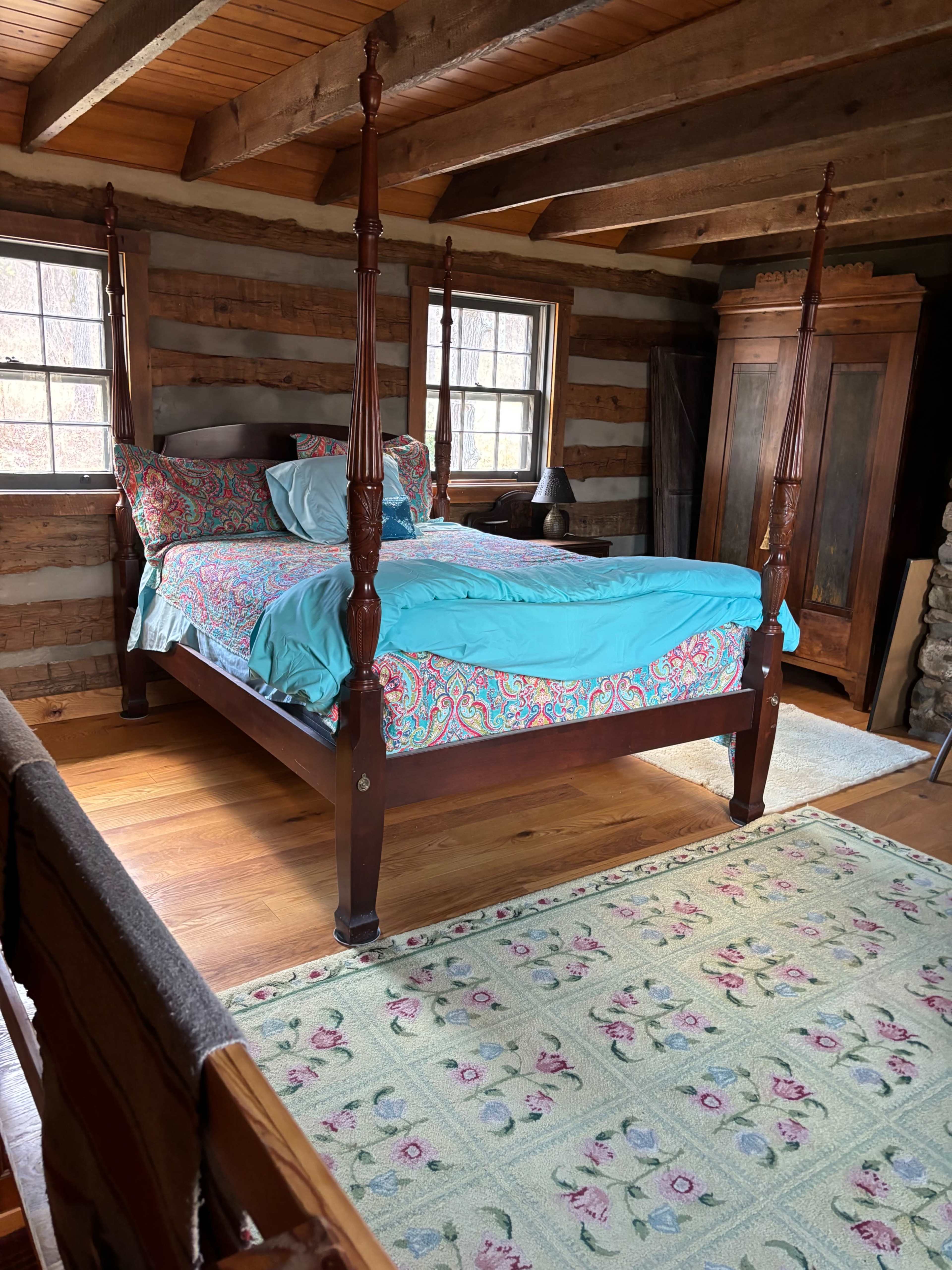 The interior of a rustic cabin features a four-poster bed with colorful bedding, hardwood floors, and exposed wooden beams.