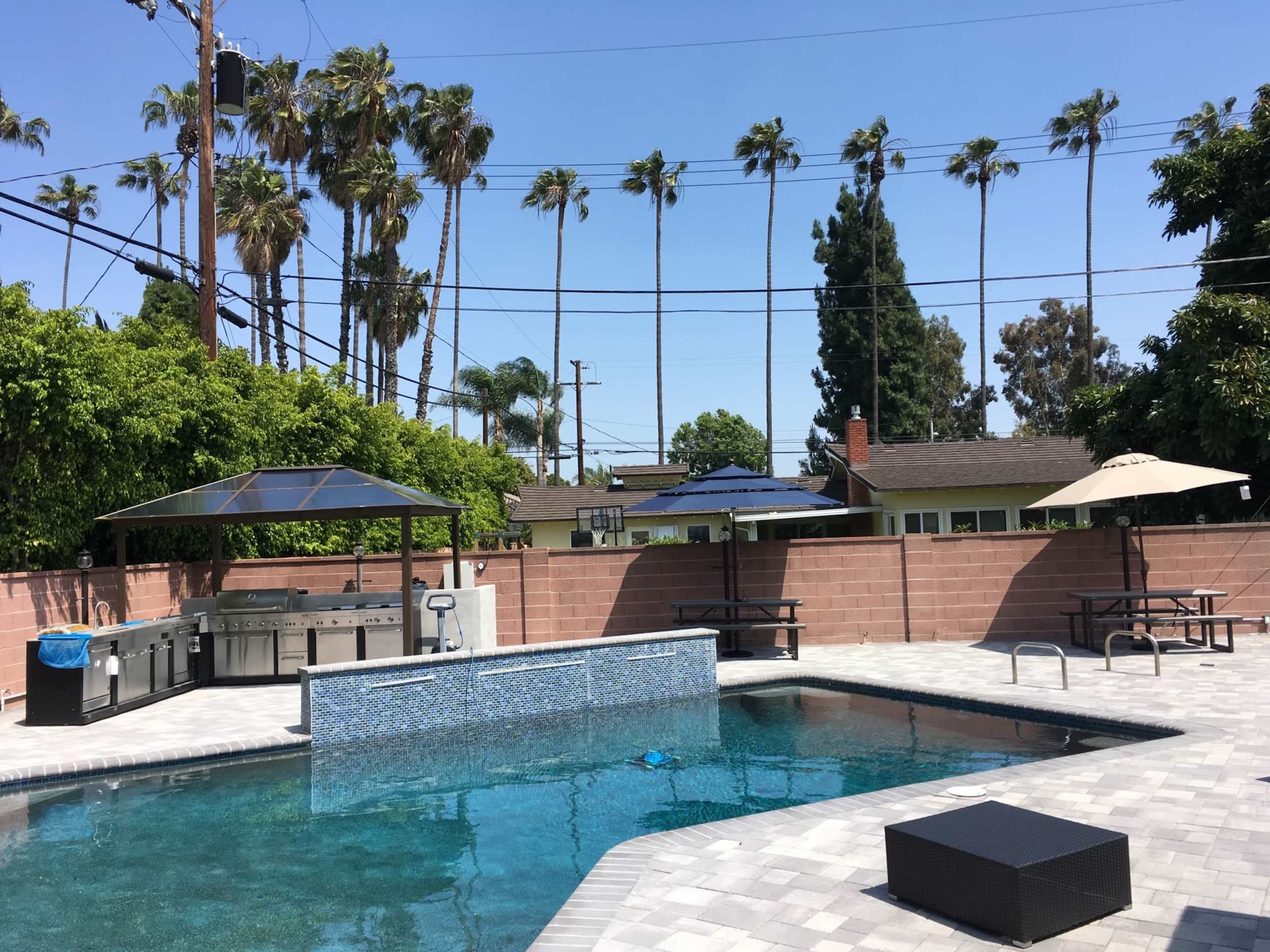 A backyard pool area features a patio with an outdoor kitchen, picnic tables, and palm trees in the background.