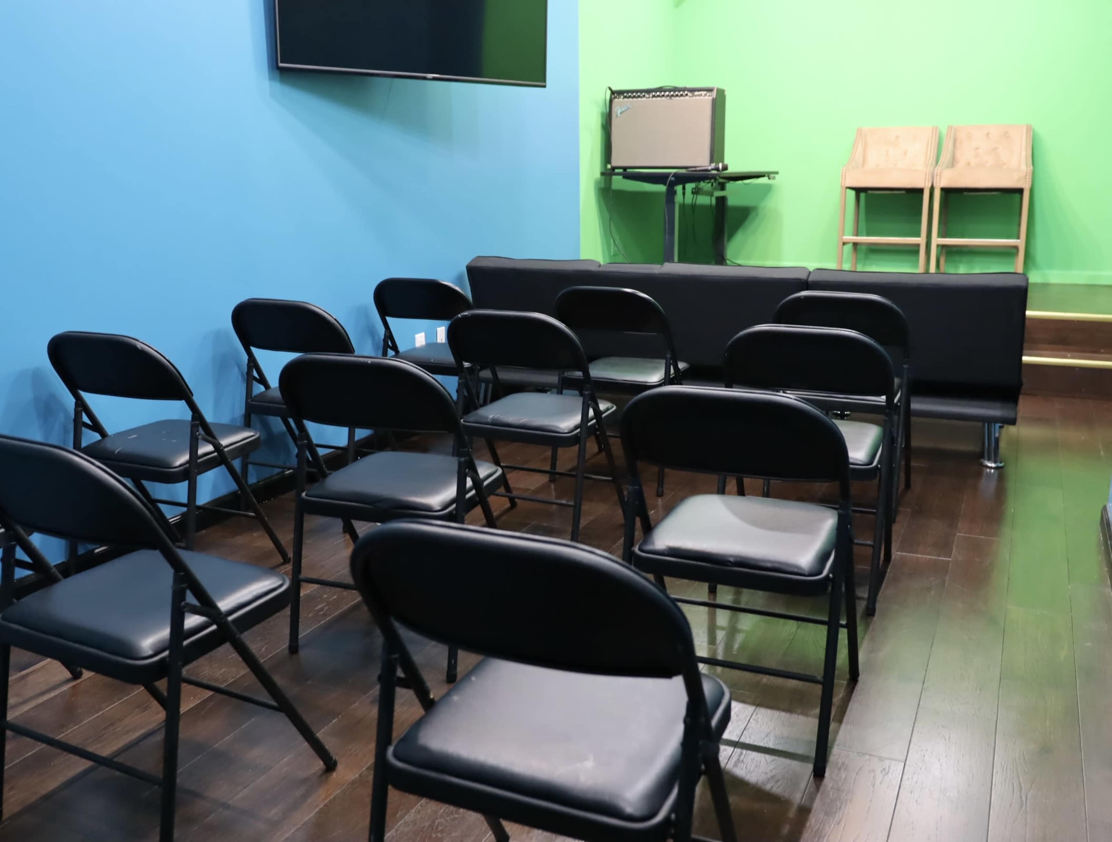 A small room with black folding chairs arranged in rows, facing a stage with an amplifier and two wooden chairs against a bright green backdrop.