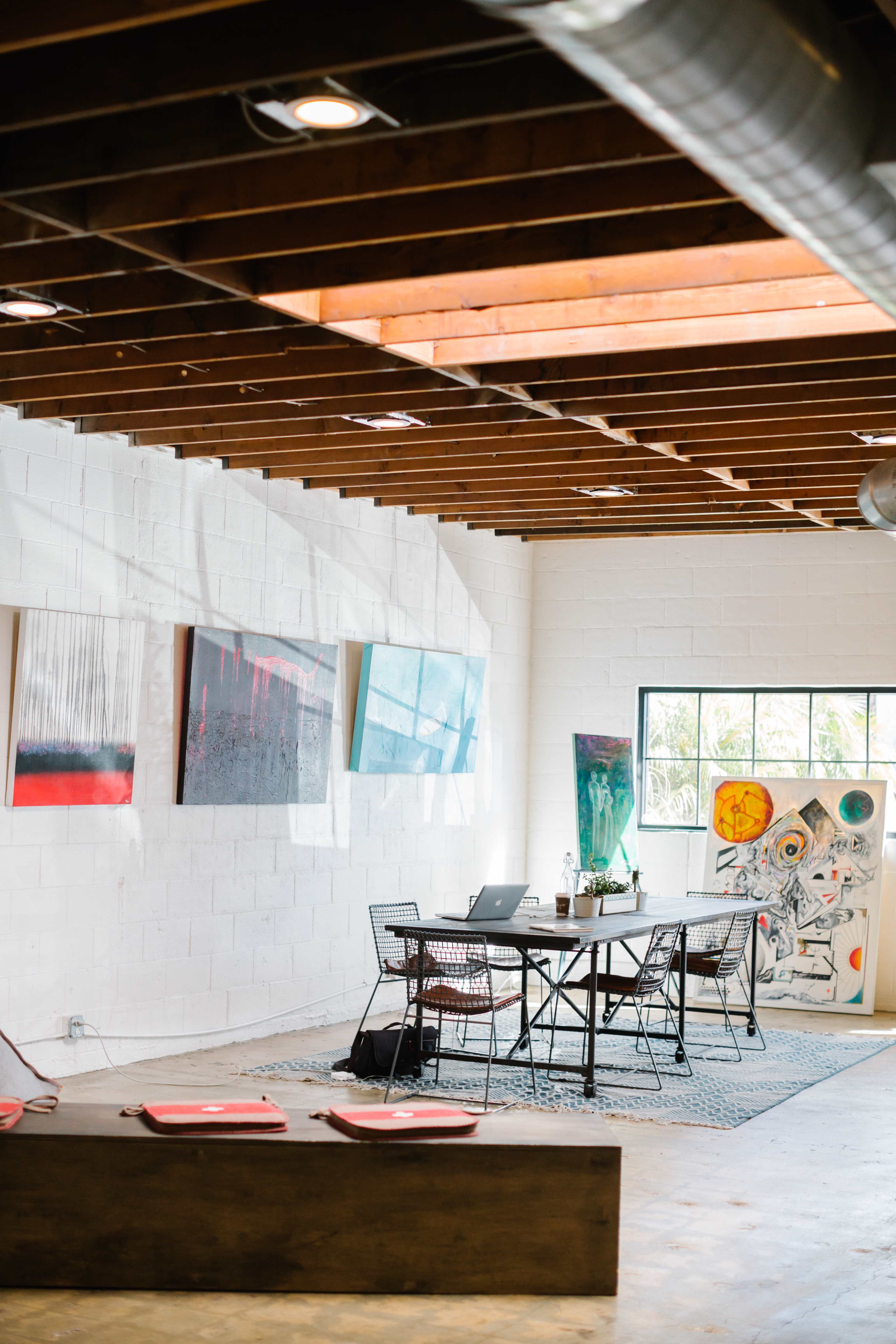 The image shows a modern workspace with a table and chairs, surrounded by various artworks on the walls and a window letting in natural light.