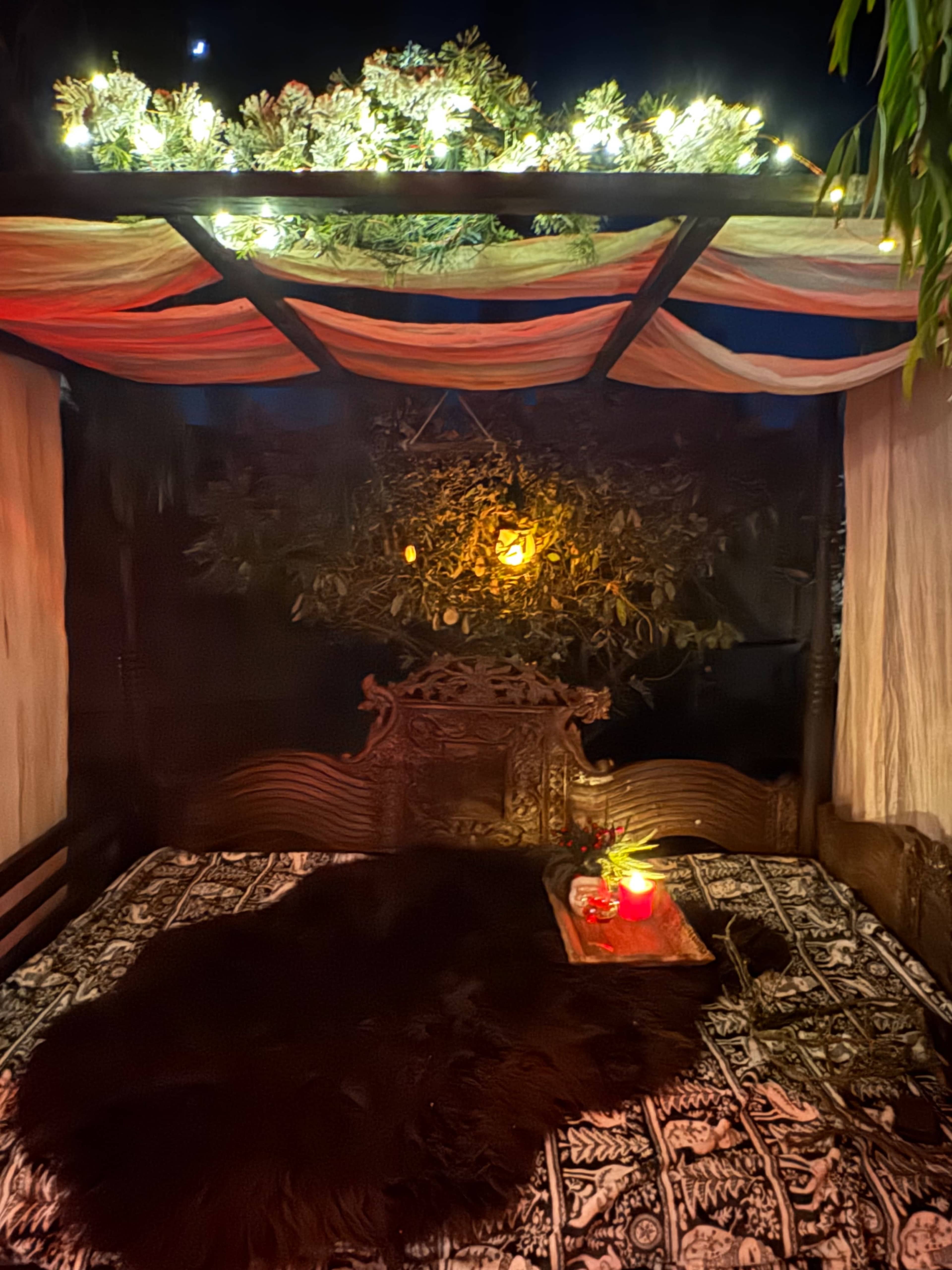The image shows a cozy outdoor seating area with a carved wooden bed, adorned with a patterned blanket, surrounded by decorative lights and greenery.