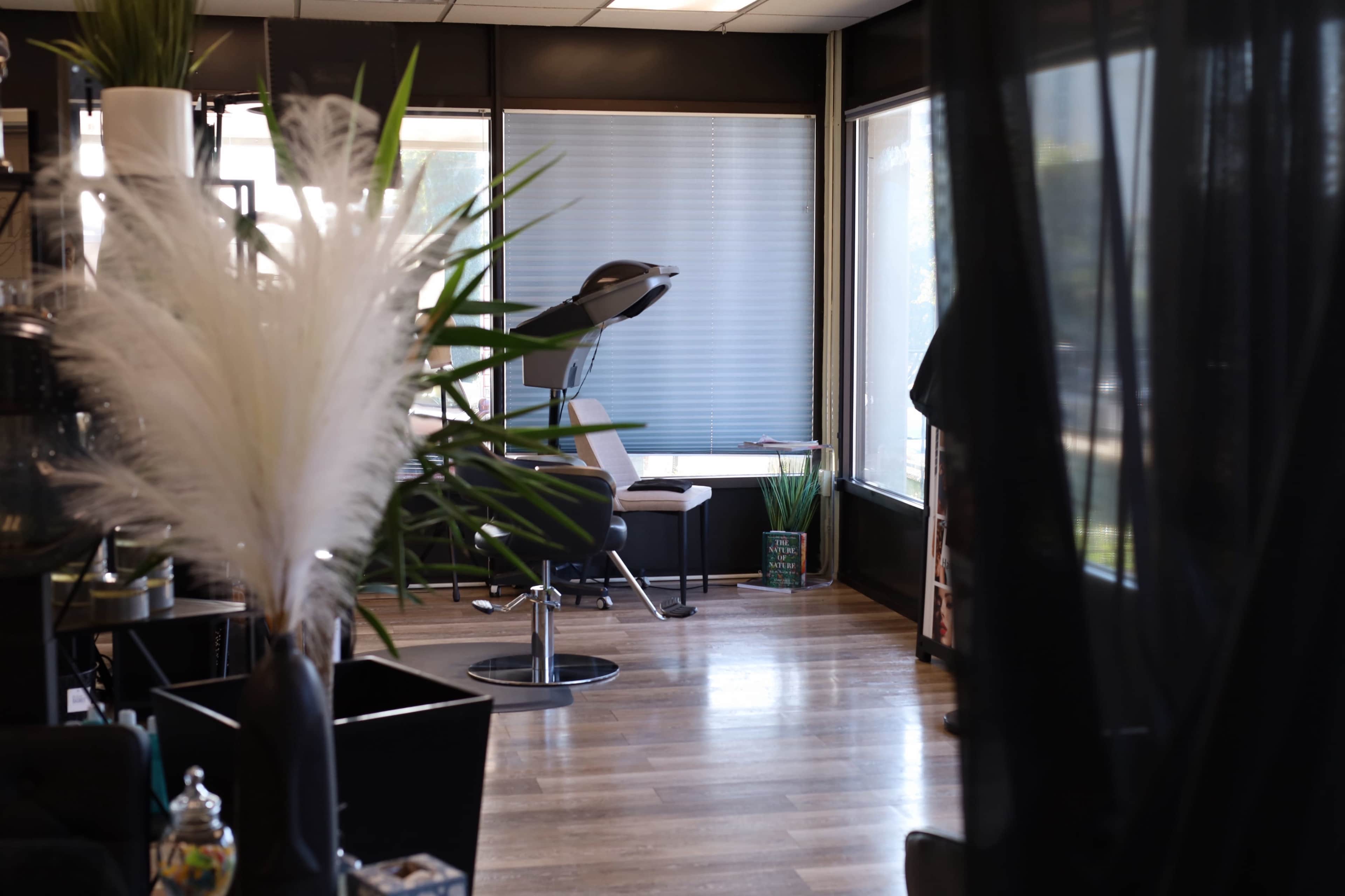 The image depicts a salon interior with a stylist chair positioned near a large window, surrounded by plants and modern furnishings.