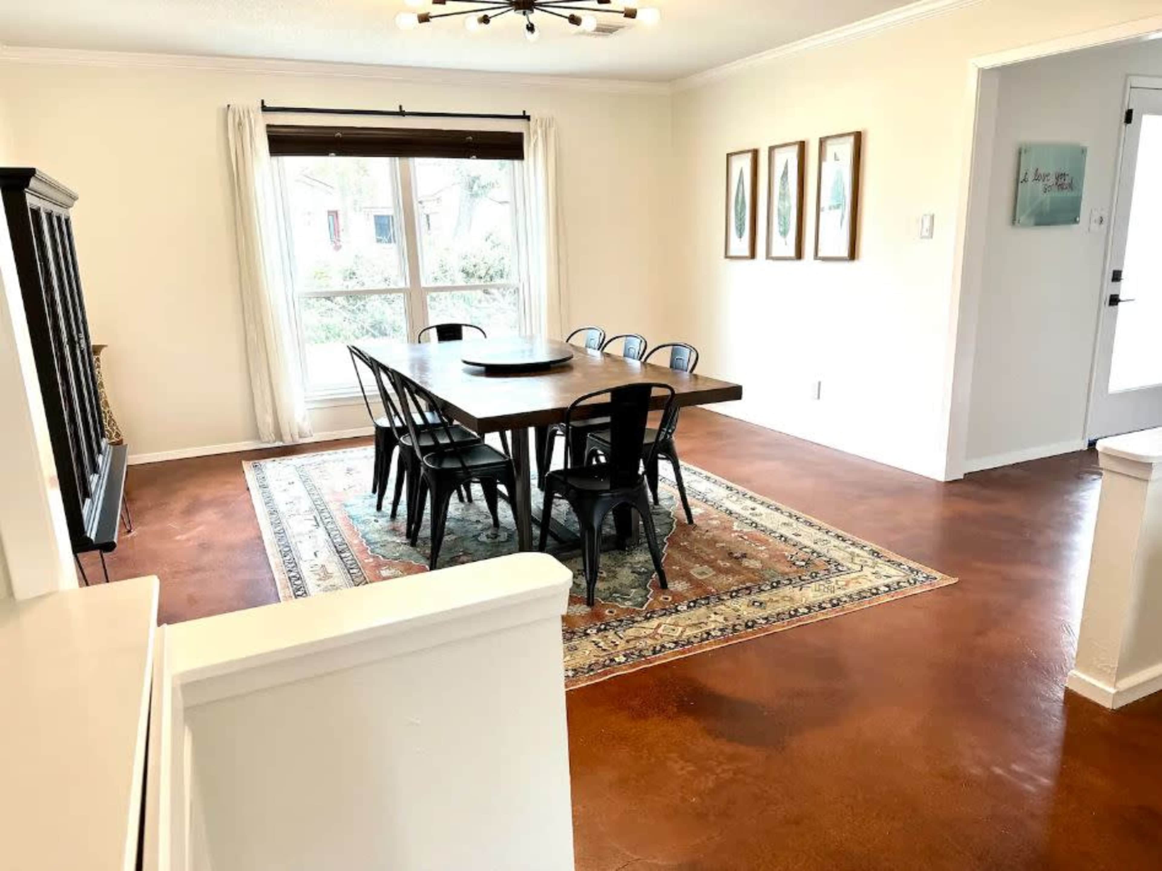 The image shows a dining area featuring a large wooden table surrounded by black chairs, a rug underneath, and framed artwork on the walls.