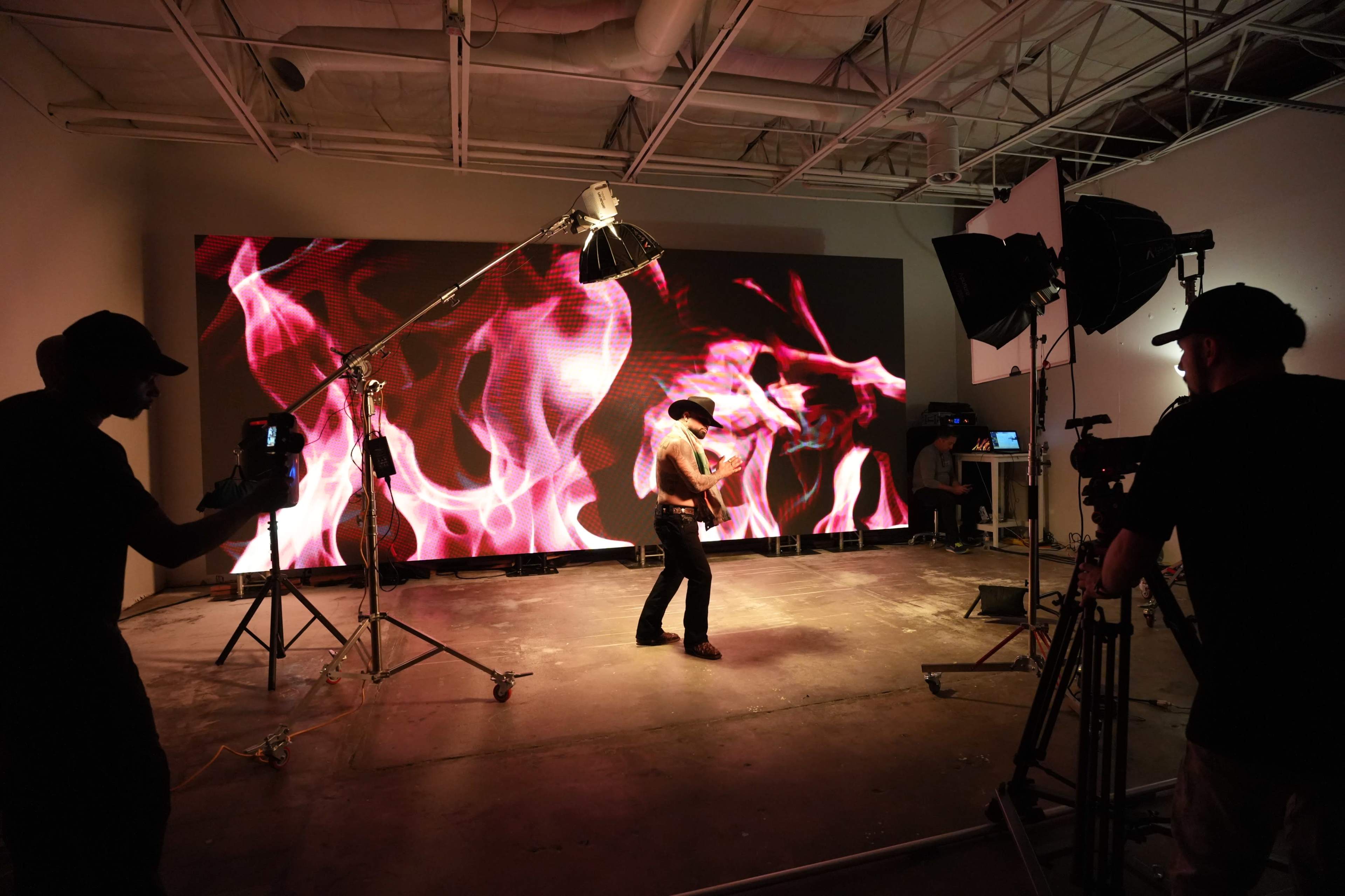 A performer is dancing on a stage in front of a large screen displaying vibrant, animated flames, while three camera operators are capturing the scene.
