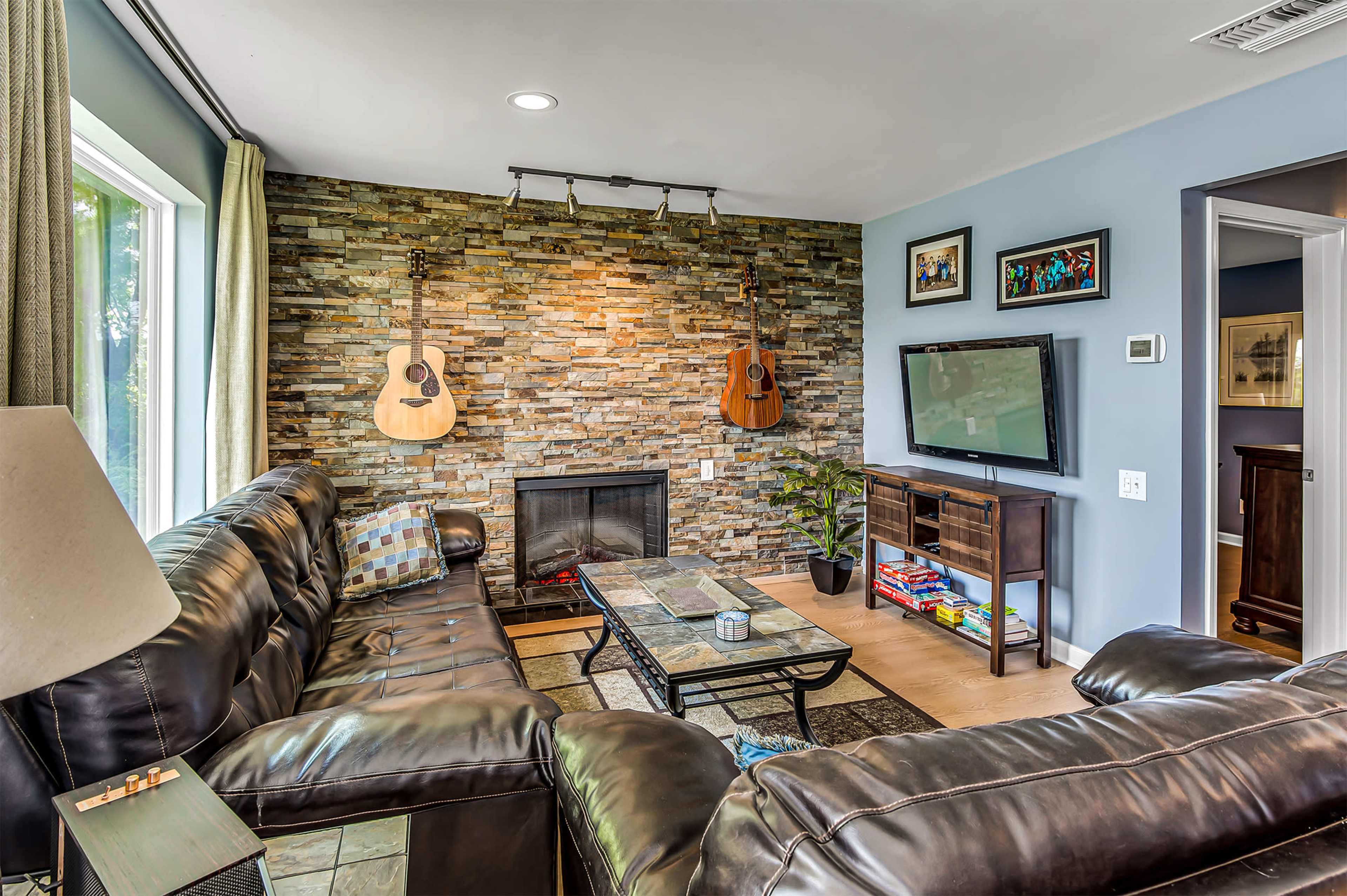 A living room with a stone accent wall featuring two guitars, a television on a stand, and a leather sectional sofa arranged around a coffee table.