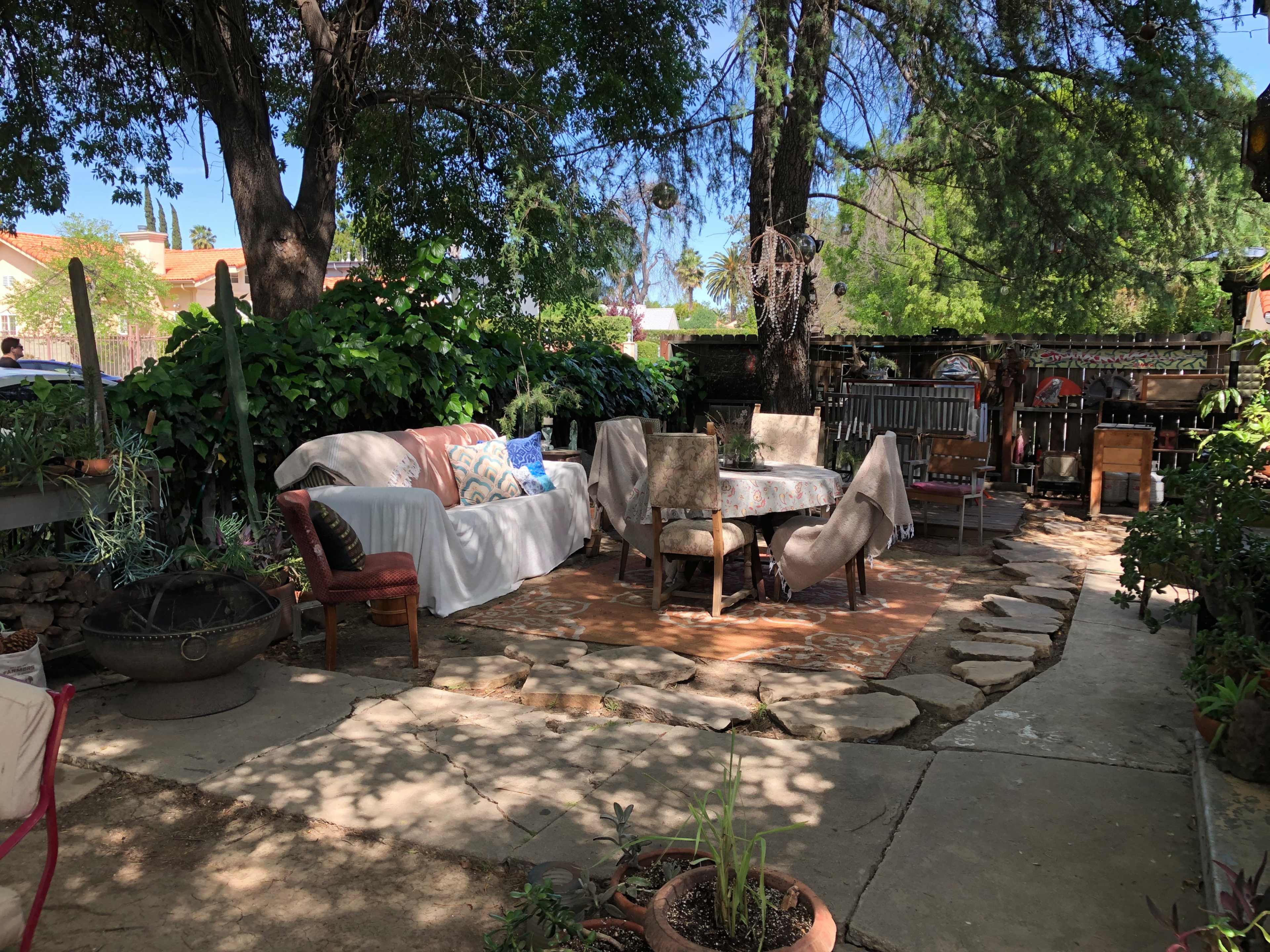 A backyard seating area with a round table surrounded by various chairs, shaded by trees and lush greenery.