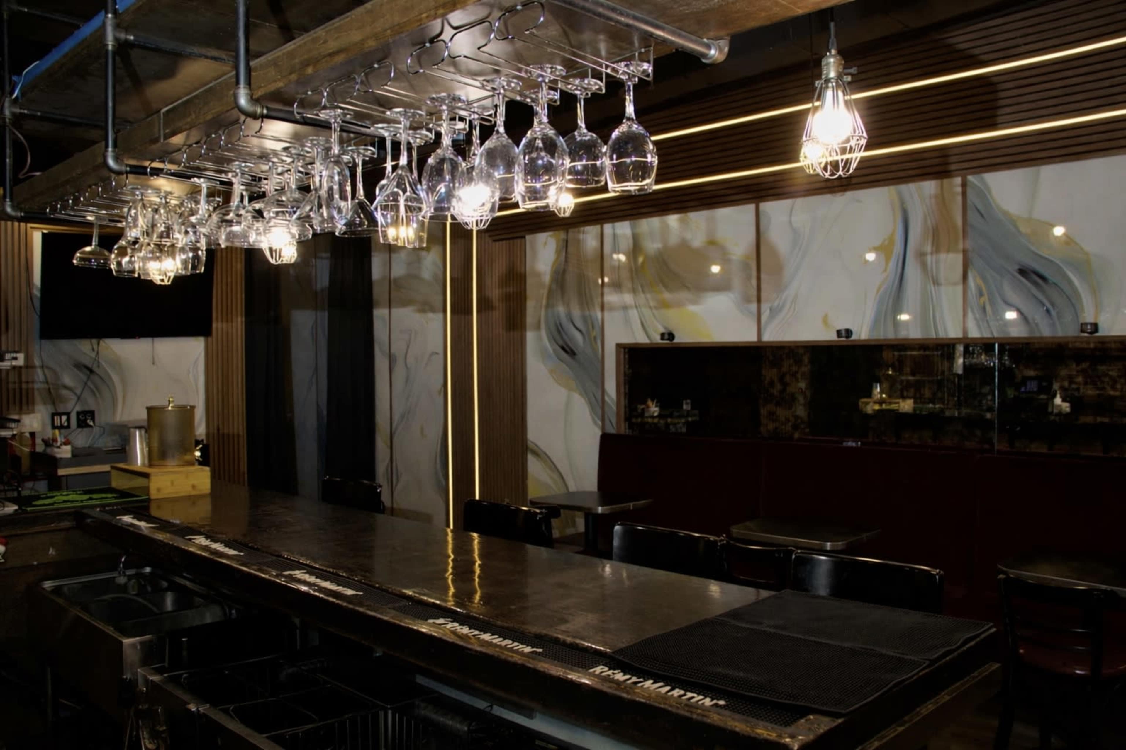 The image shows a bar area with a metal counter, hanging glasses, and warm lighting, featuring a minimalist design and a backdrop of decorative wall patterns.