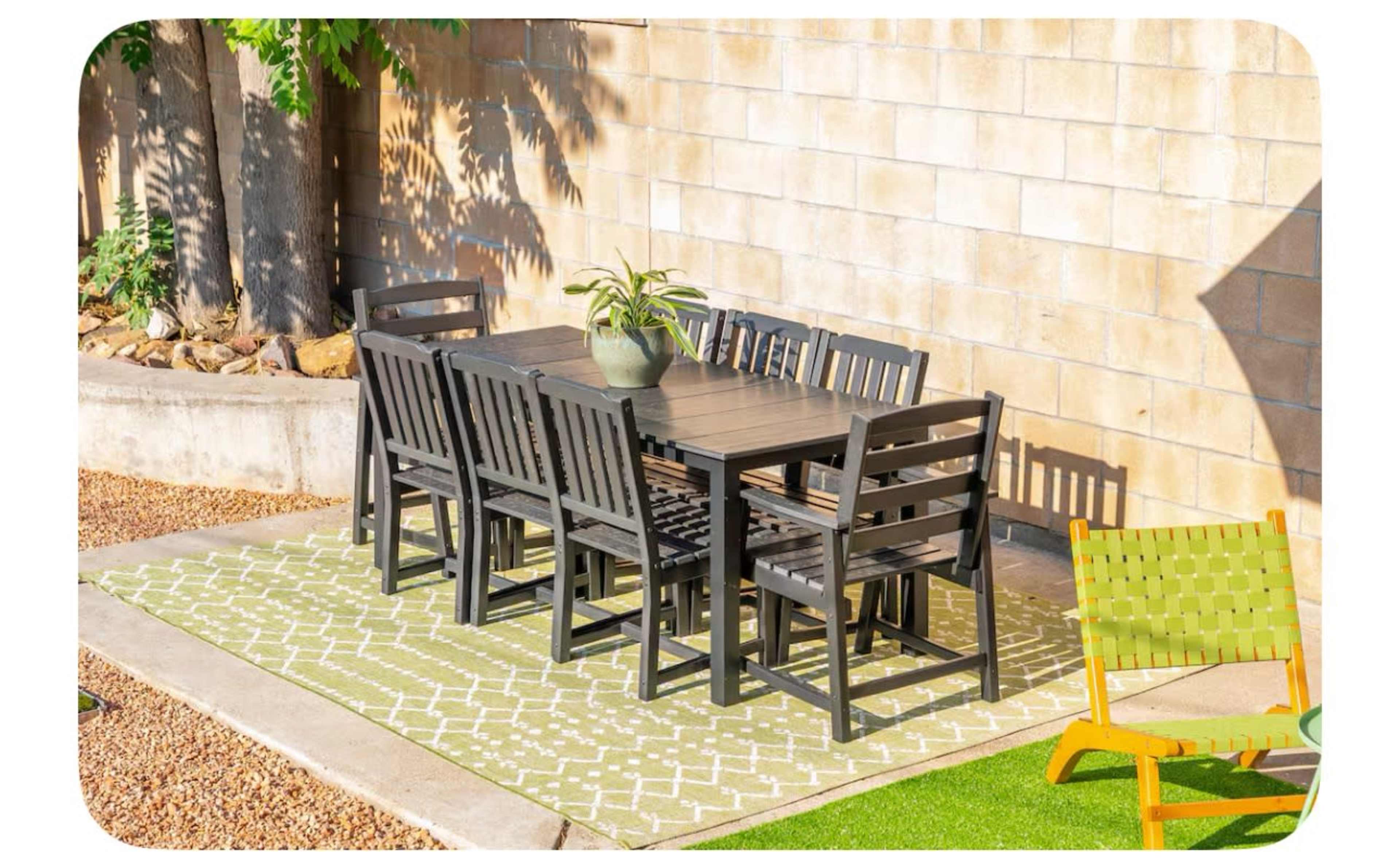 A rectangular black dining table with eight matching chairs is set on a green patterned rug against a textured wall in an outdoor patio area.