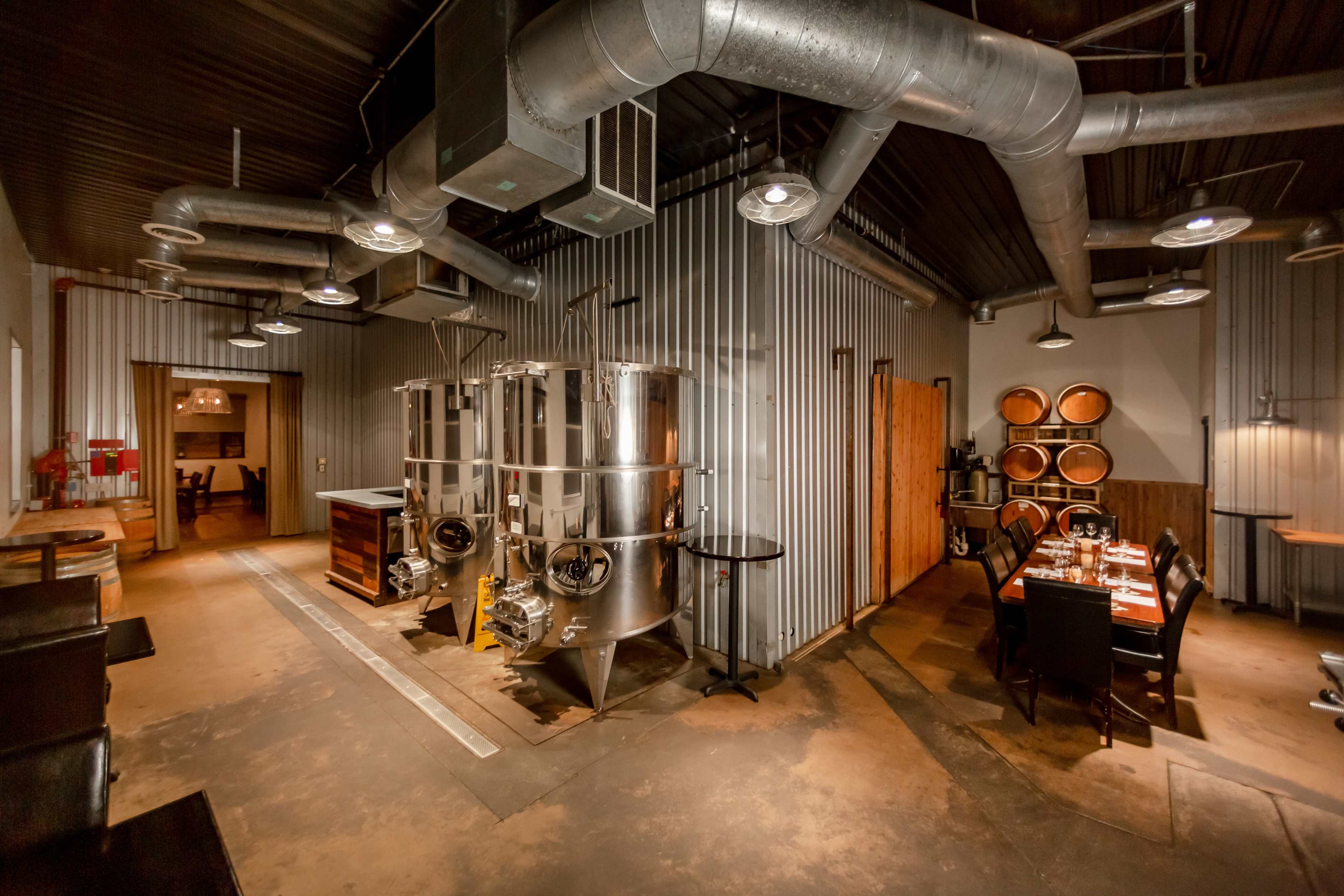 The image shows an industrial-style interior featuring metal fermentation tanks, wooden barrels, and a dining area with a table set for multiple guests.