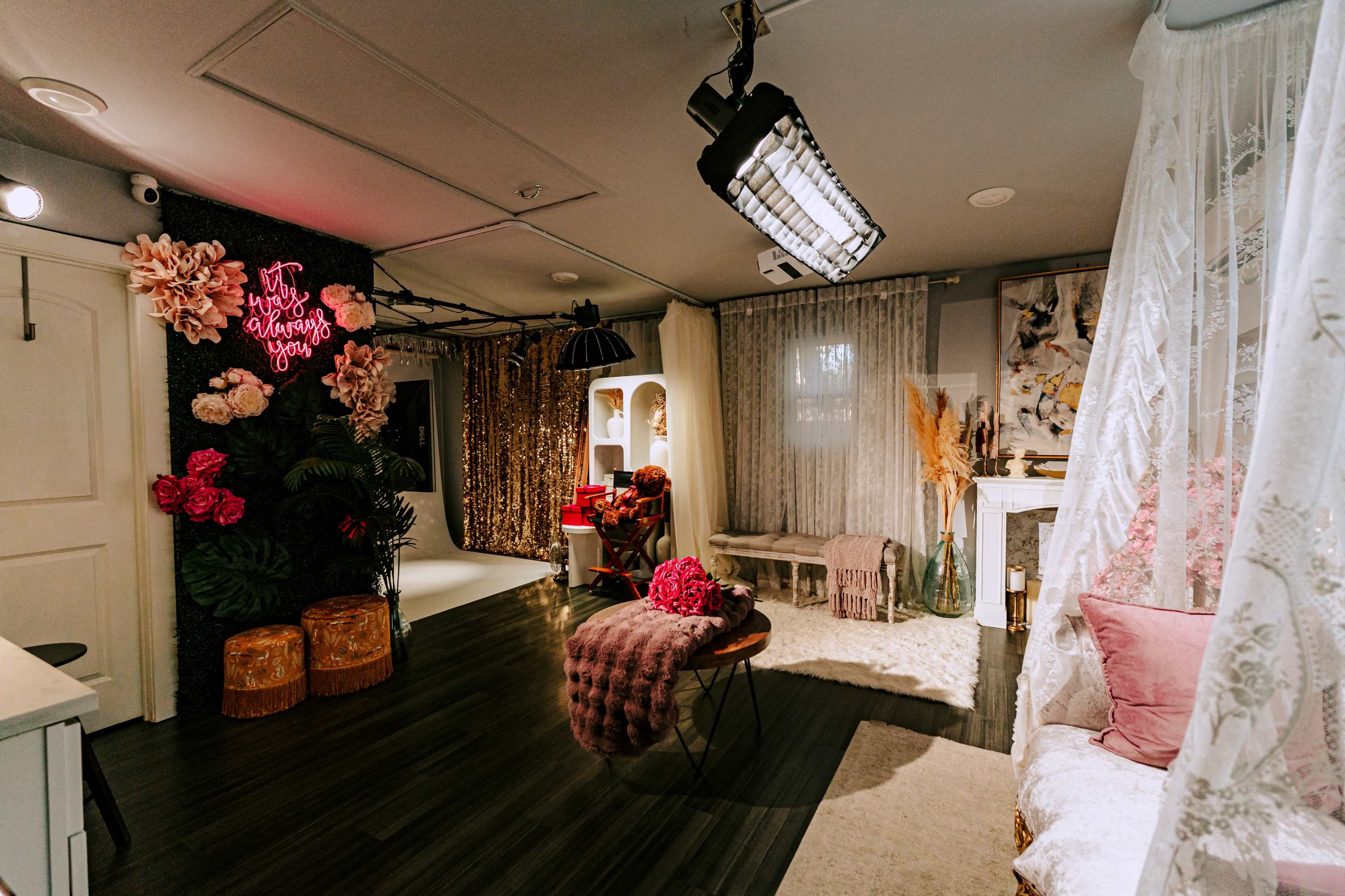 The image shows a beautifully decorated room featuring floral accents, a cozy seating area, and a bright neon sign, creating a vibrant and inviting atmosphere.