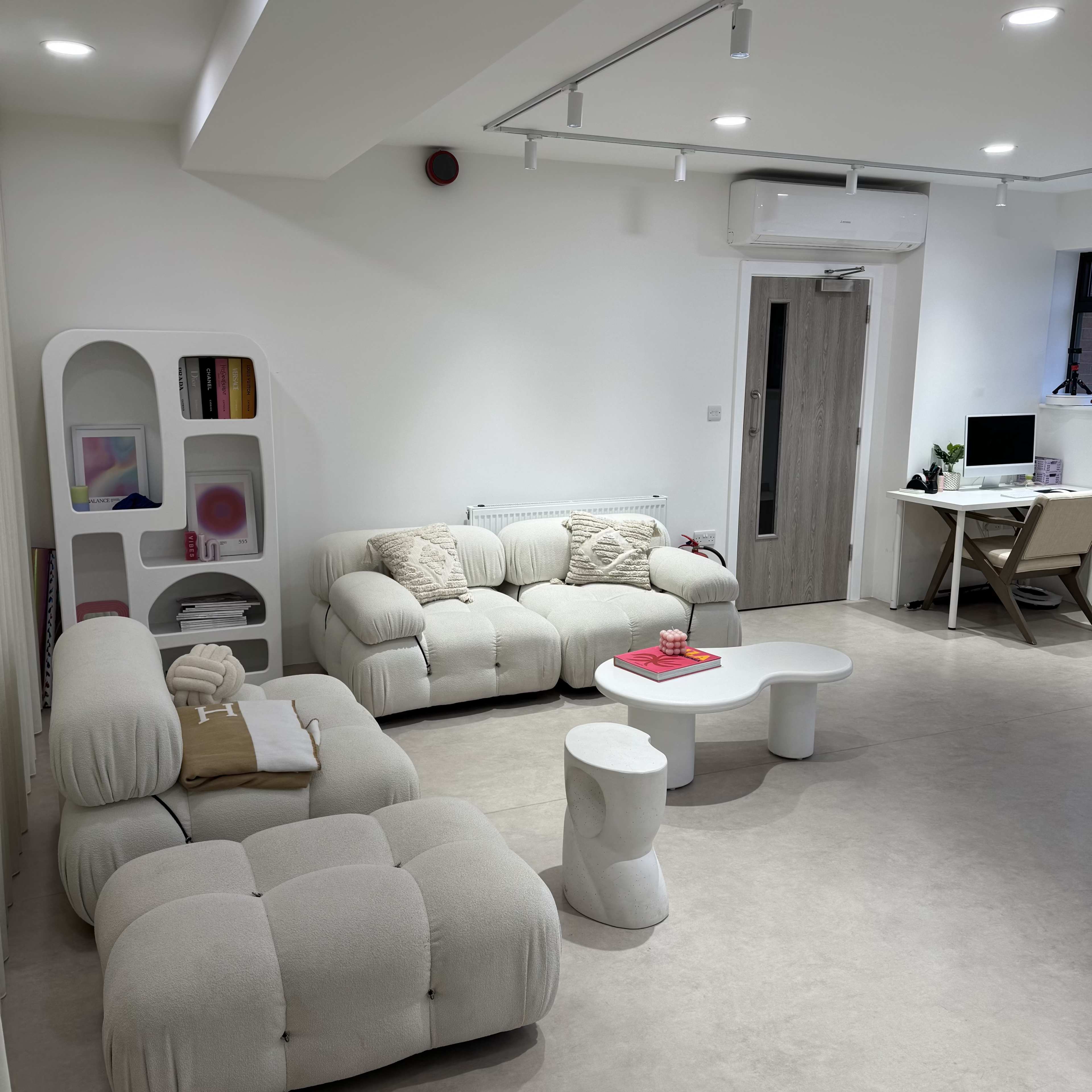The image shows a modern, minimalist living room with two beige sofas, a white coffee table, and a desk with a computer in the corner.