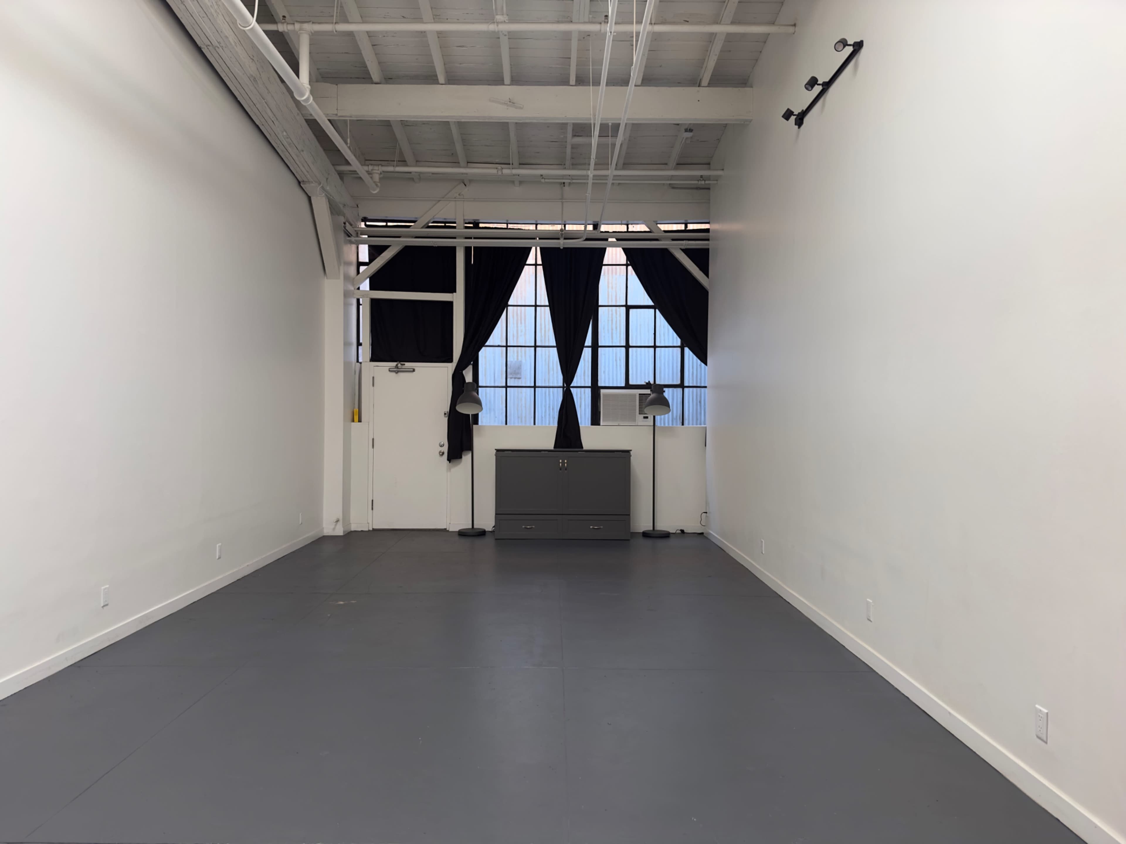 The image shows a spacious, empty room with white walls, a concrete floor, and large windows covered by black curtains.