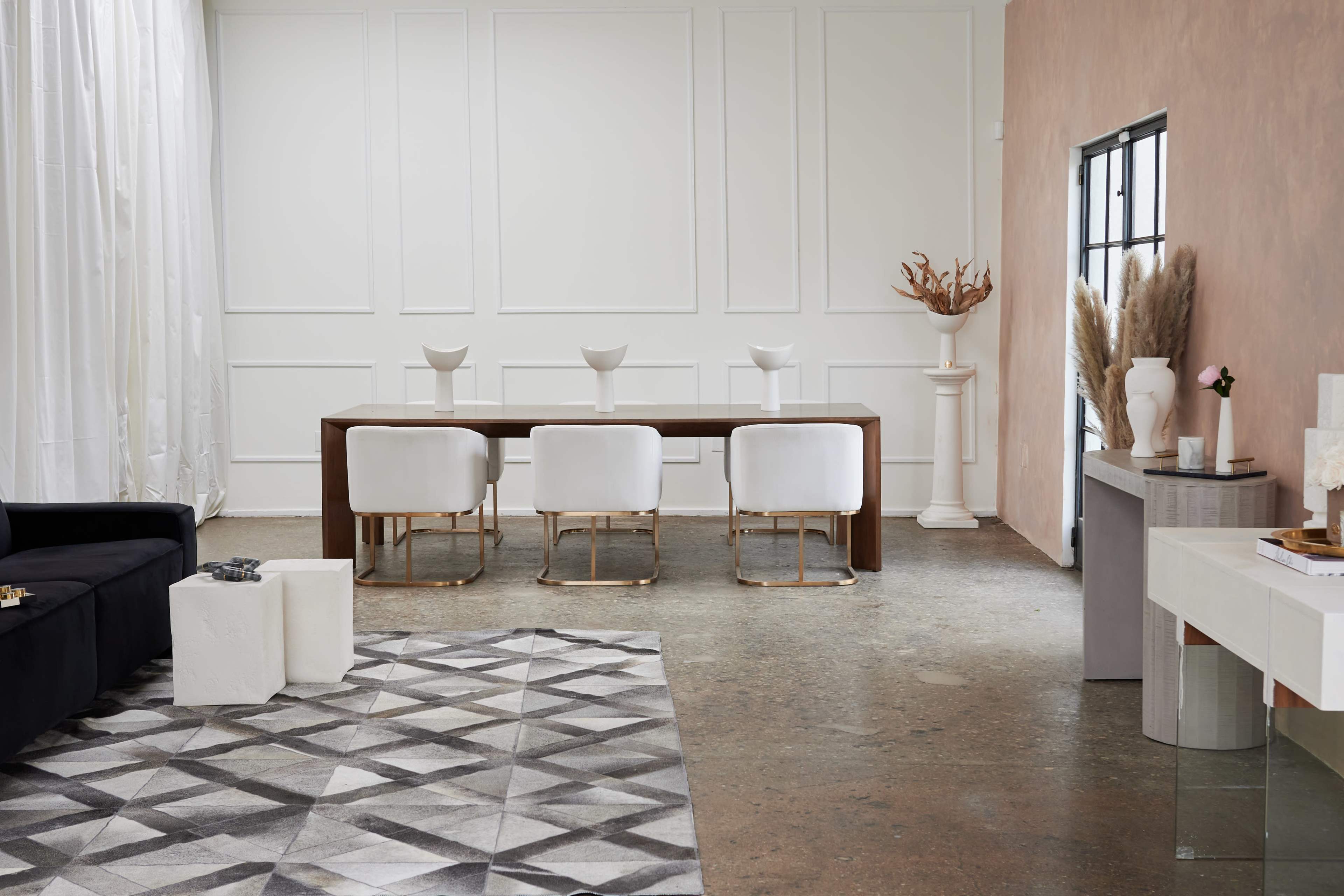 A modern dining area features a long wooden table surrounded by white chairs, with a geometric rug on the floor and minimalist decor along the walls.