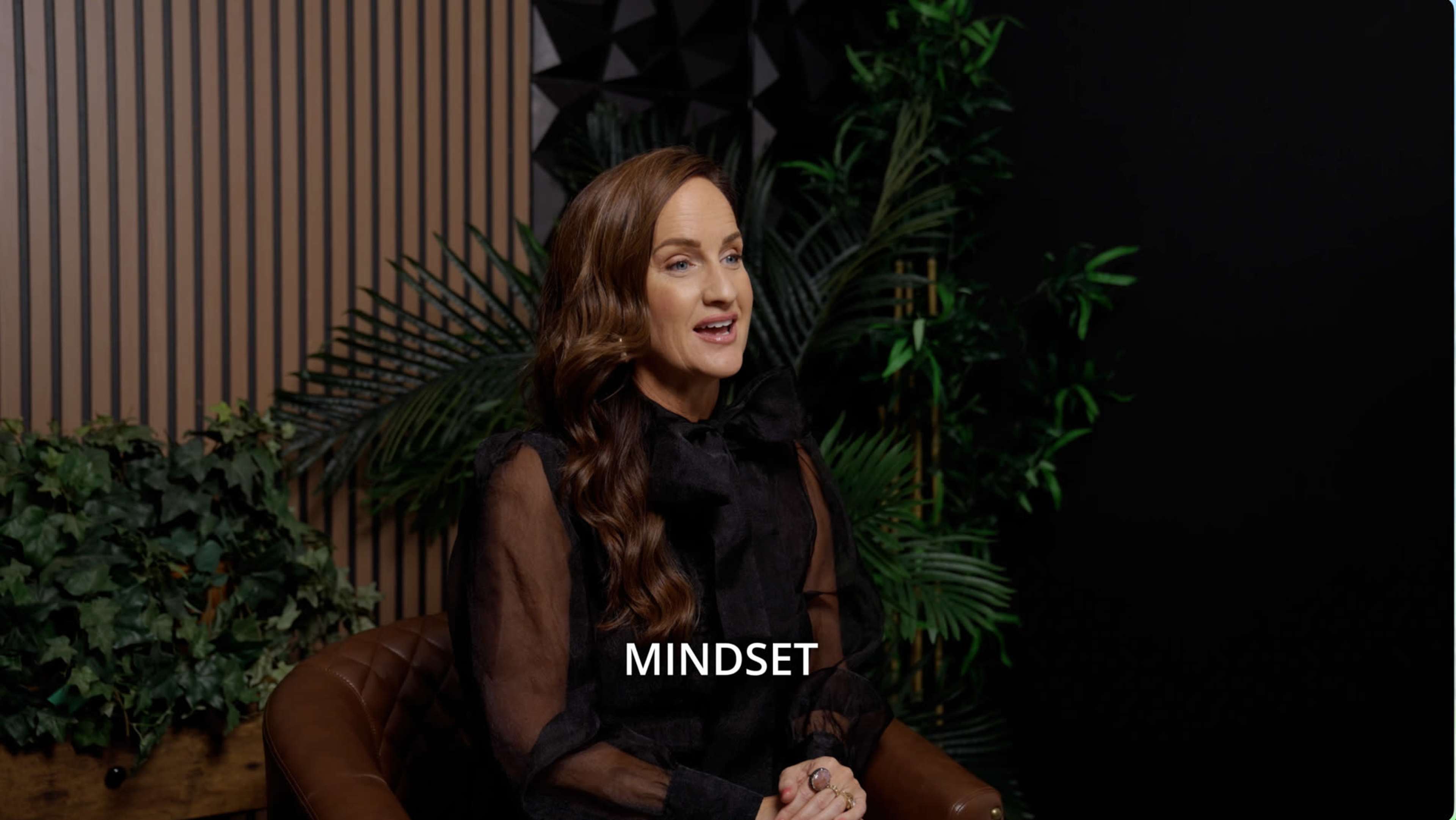 A woman with long hair sits in a chair, speaking in front of a backdrop featuring plants and textured wall panels, with the word "MINDSET" displayed below her.