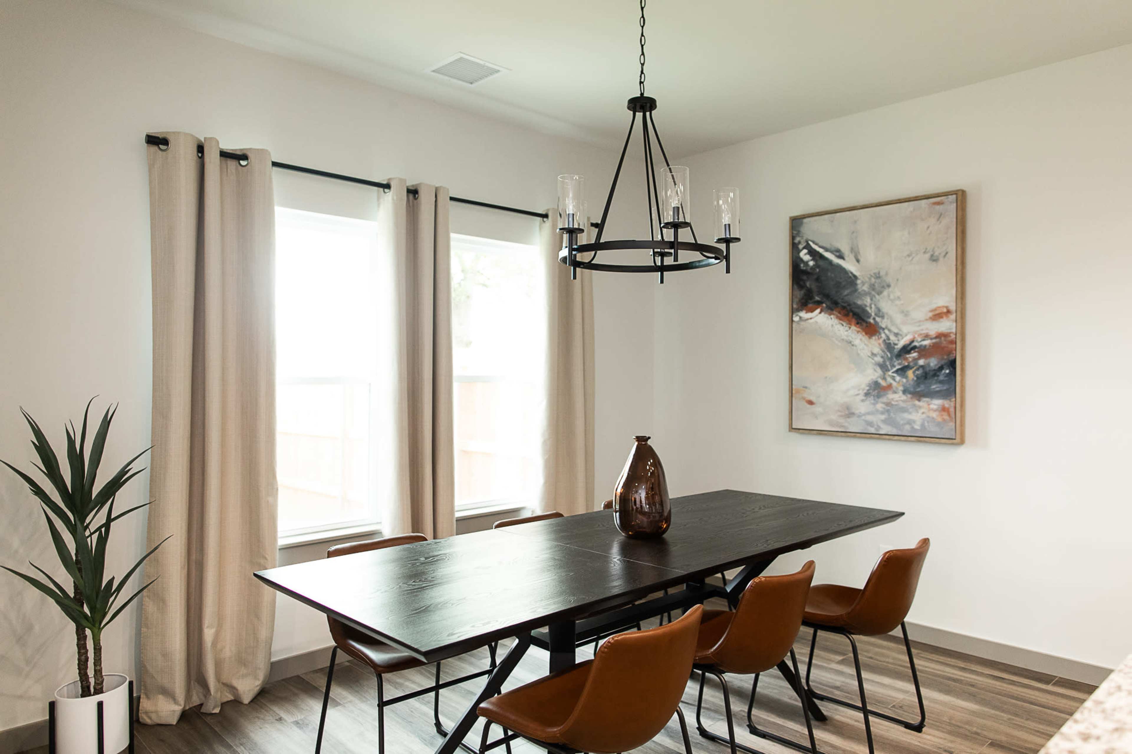 A modern dining room features a long black table surrounded by brown chairs, with large windows and a contemporary chandelier overhead.