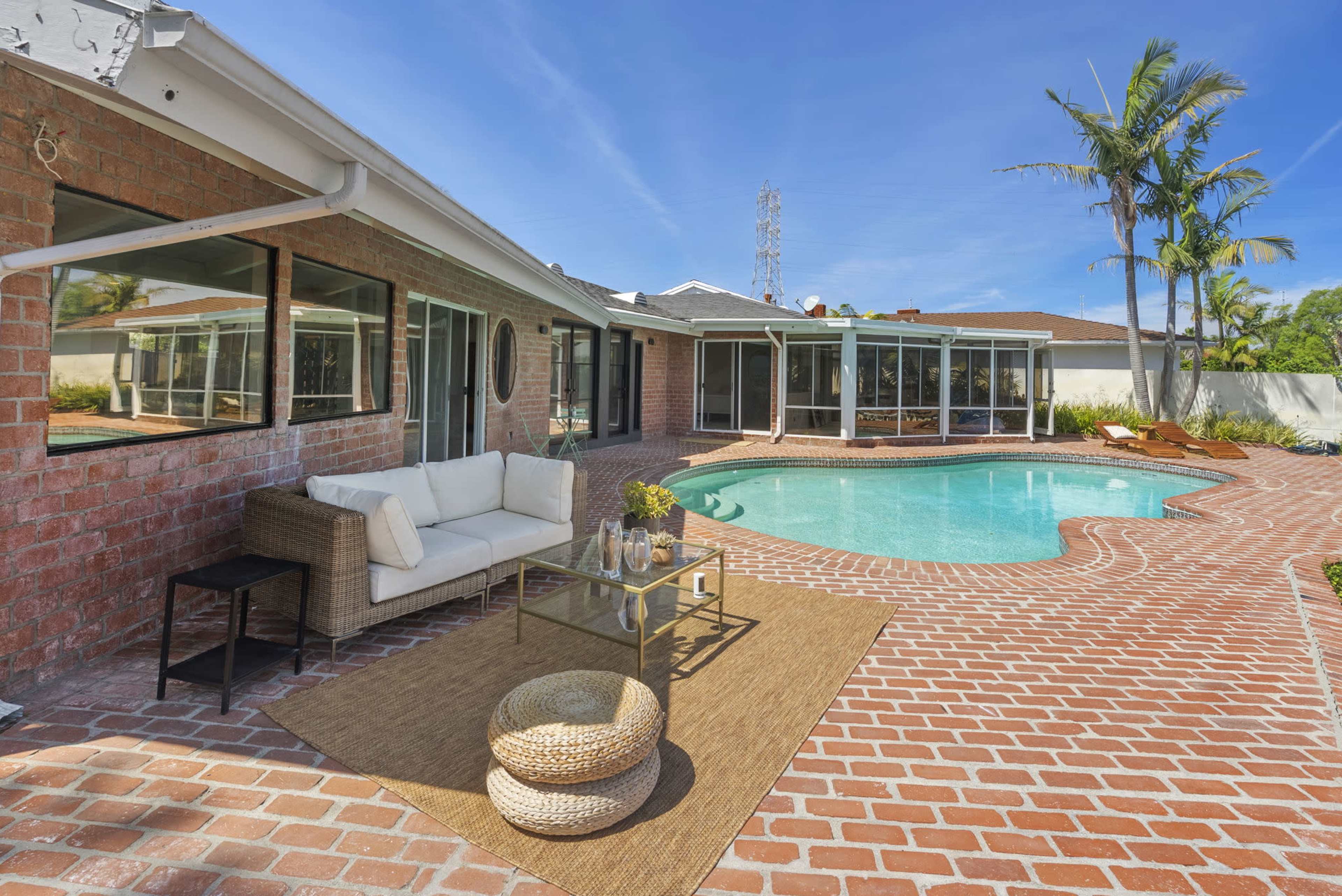 The image shows a backyard with a brick patio, a swimming pool, and a seating area featuring a white couch and a glass coffee table, surrounded by palm trees.