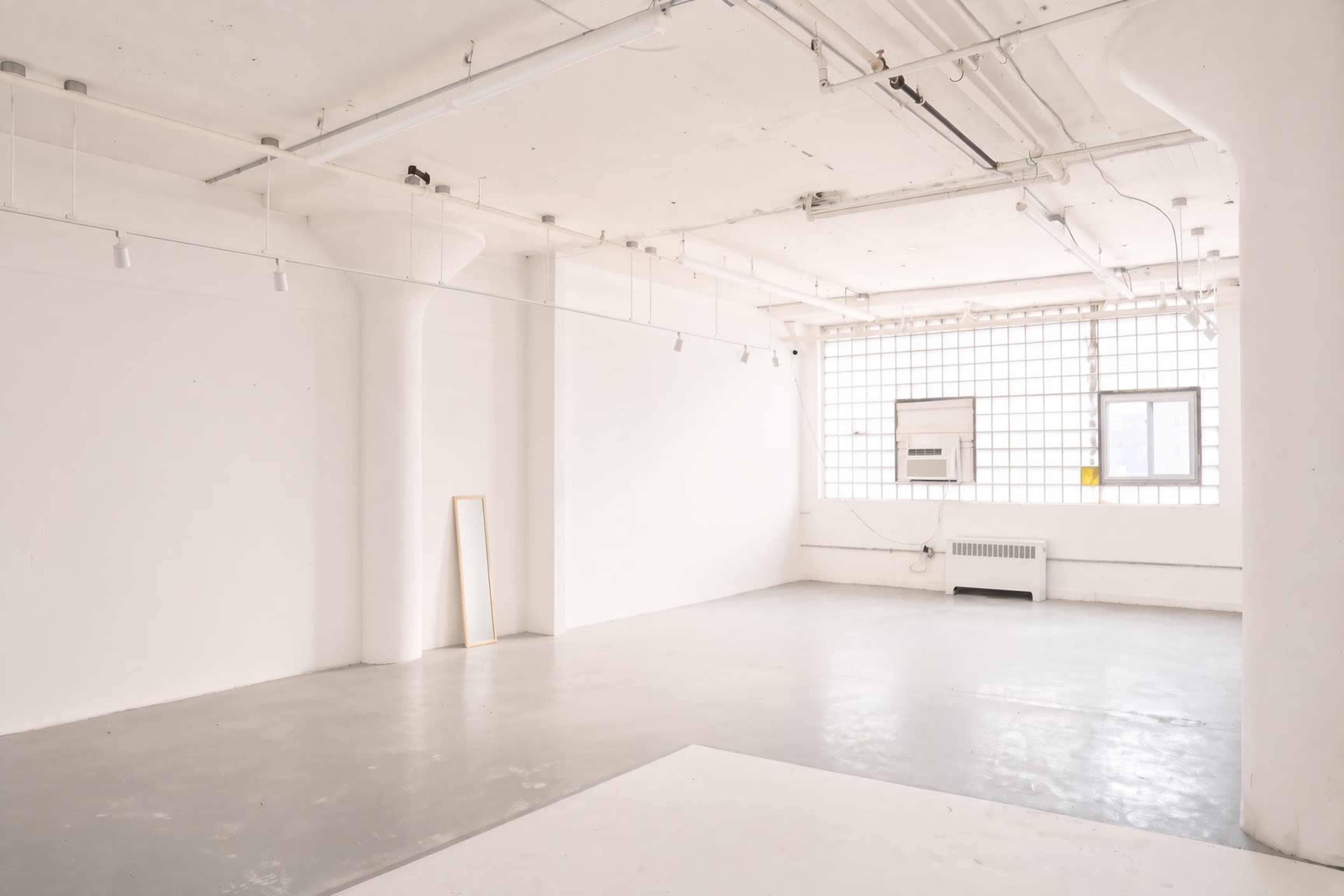 The image depicts a spacious, empty room with white walls, concrete flooring, and large windows allowing natural light to enter.