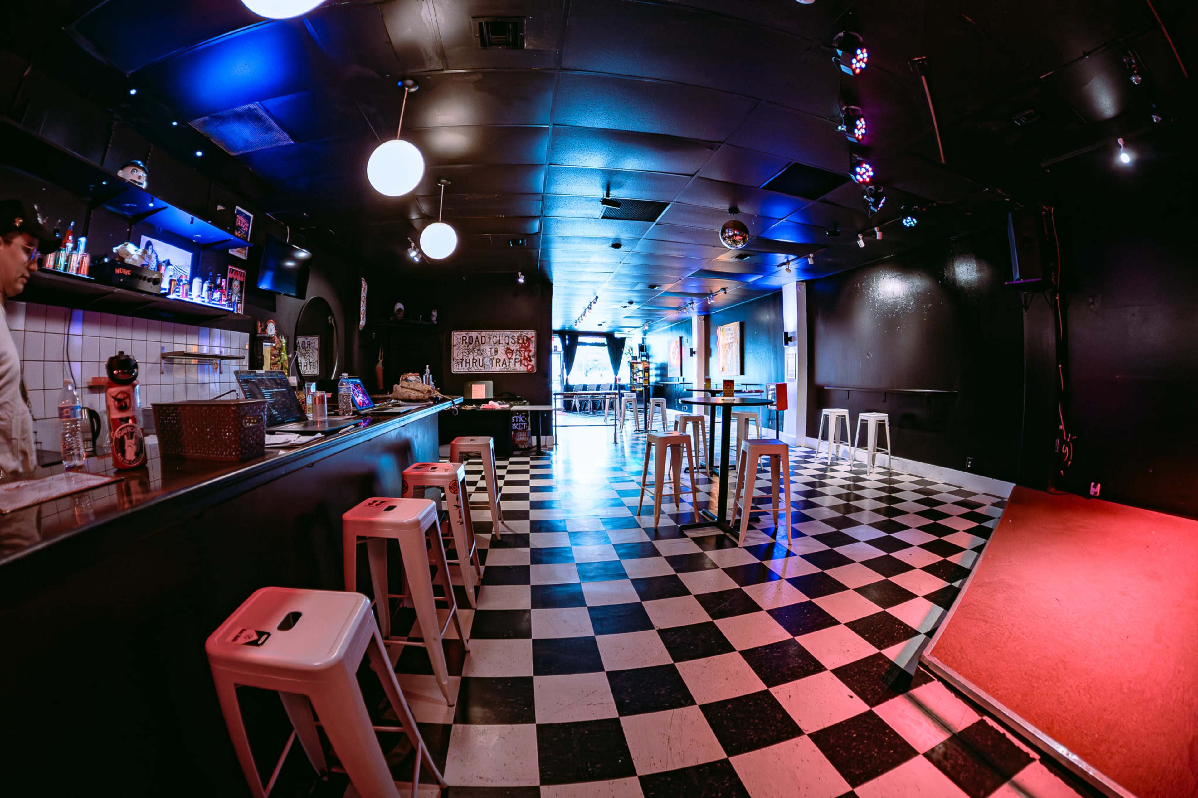 The image shows a dimly lit bar with a checkered floor, illuminated by colorful lights, featuring a long counter on one side and a small stage area.