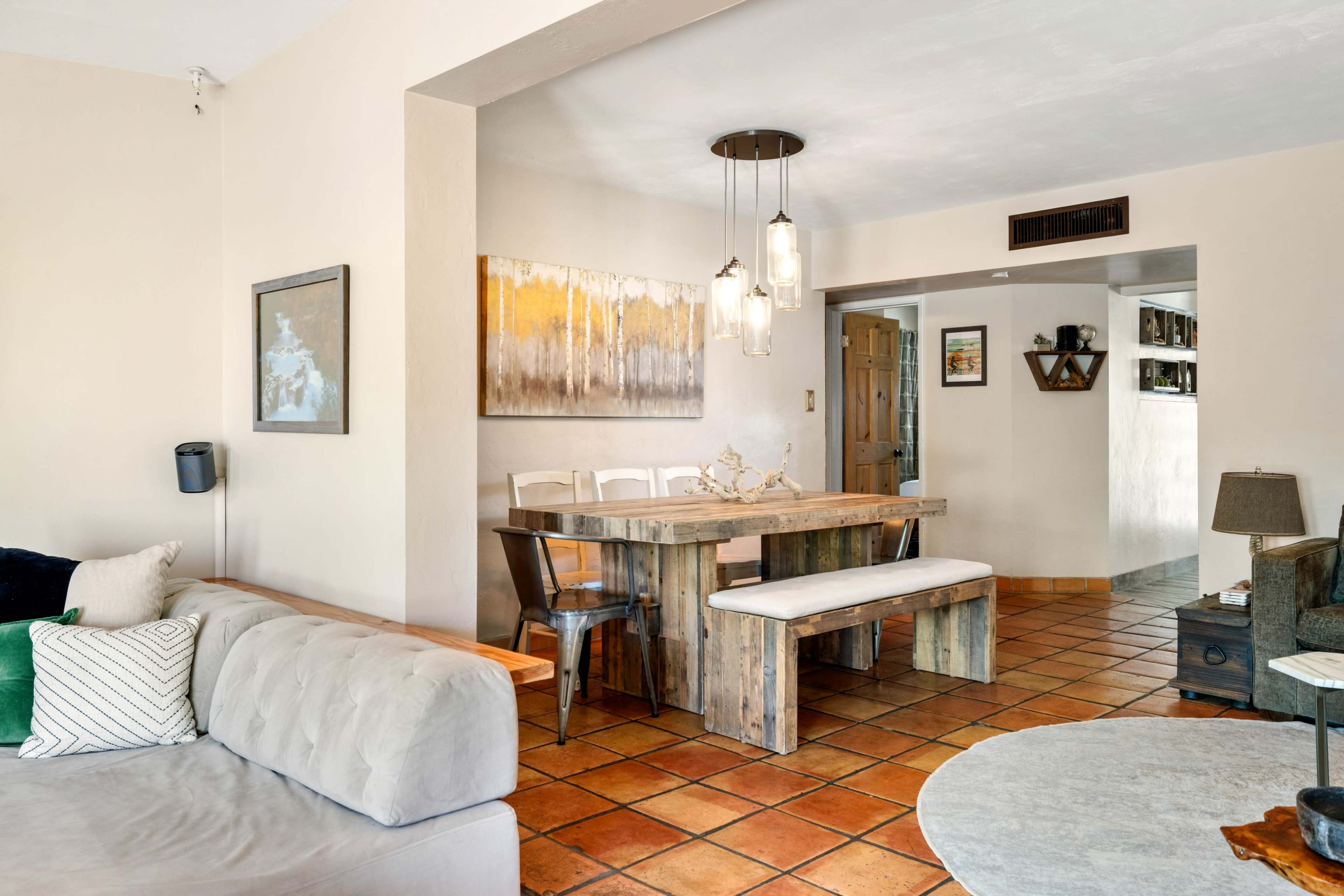The image shows a bright, open living space featuring a dining area with a wooden table and benches, adjacent to a cozy seating area with a sofa.