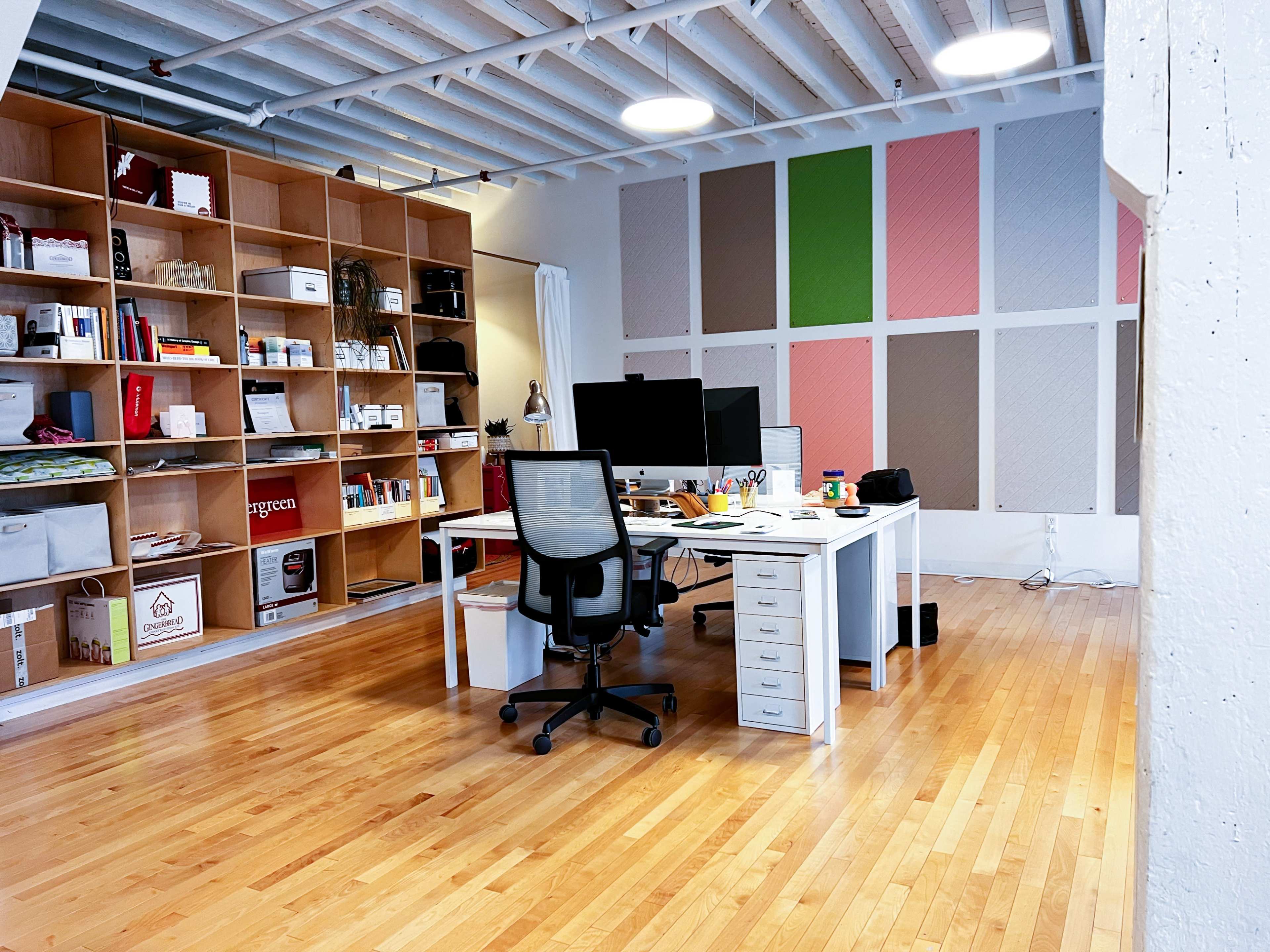 A bright, modern office space features a desk with two computer monitors, a filled bookshelf, and colorful wall panels.
