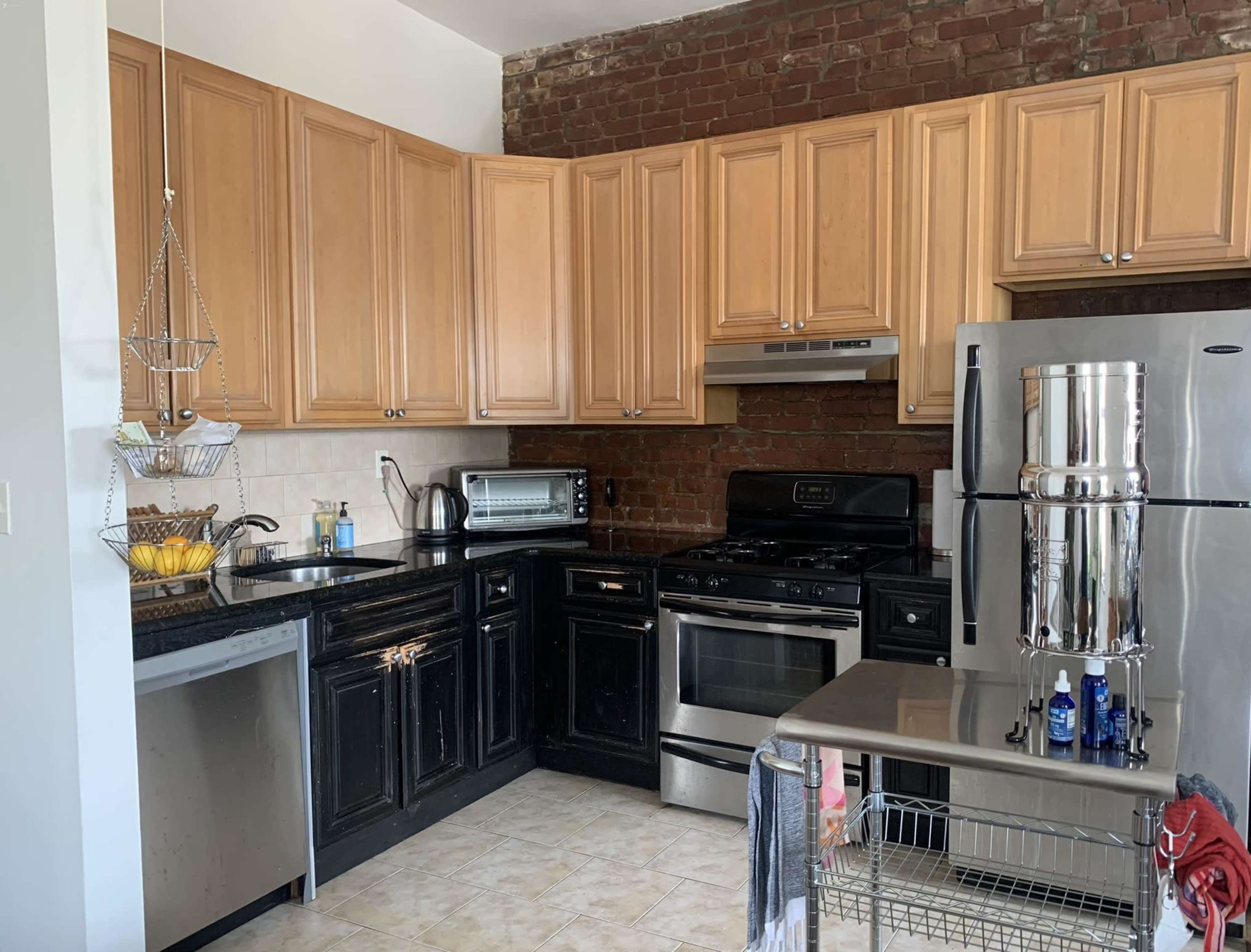 A kitchen features wooden cabinets, a black countertop, stainless steel appliances, and exposed brick walls.