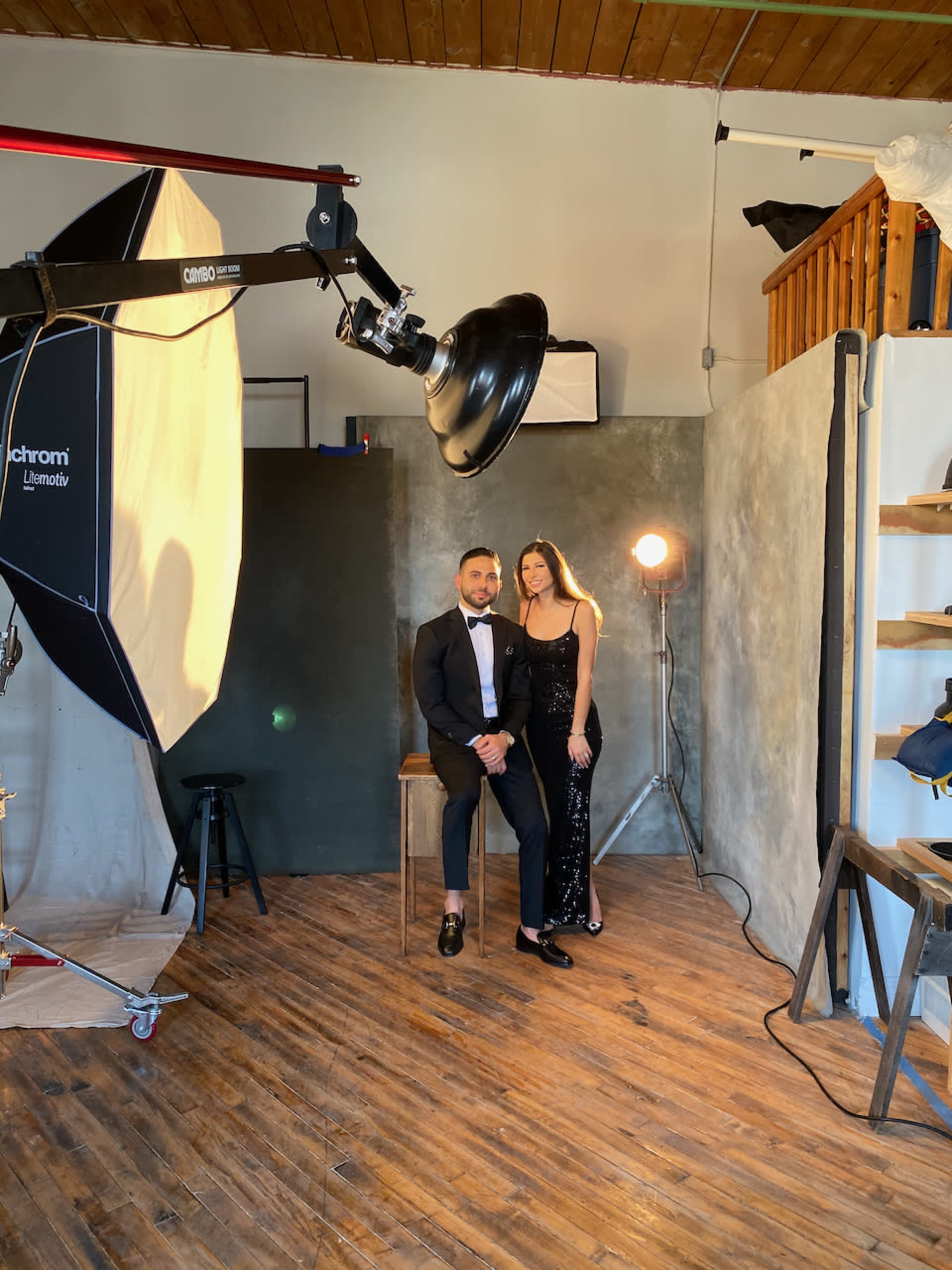 A man in a tuxedo and a woman in a black dress pose together in a photography studio with professional lighting equipment.