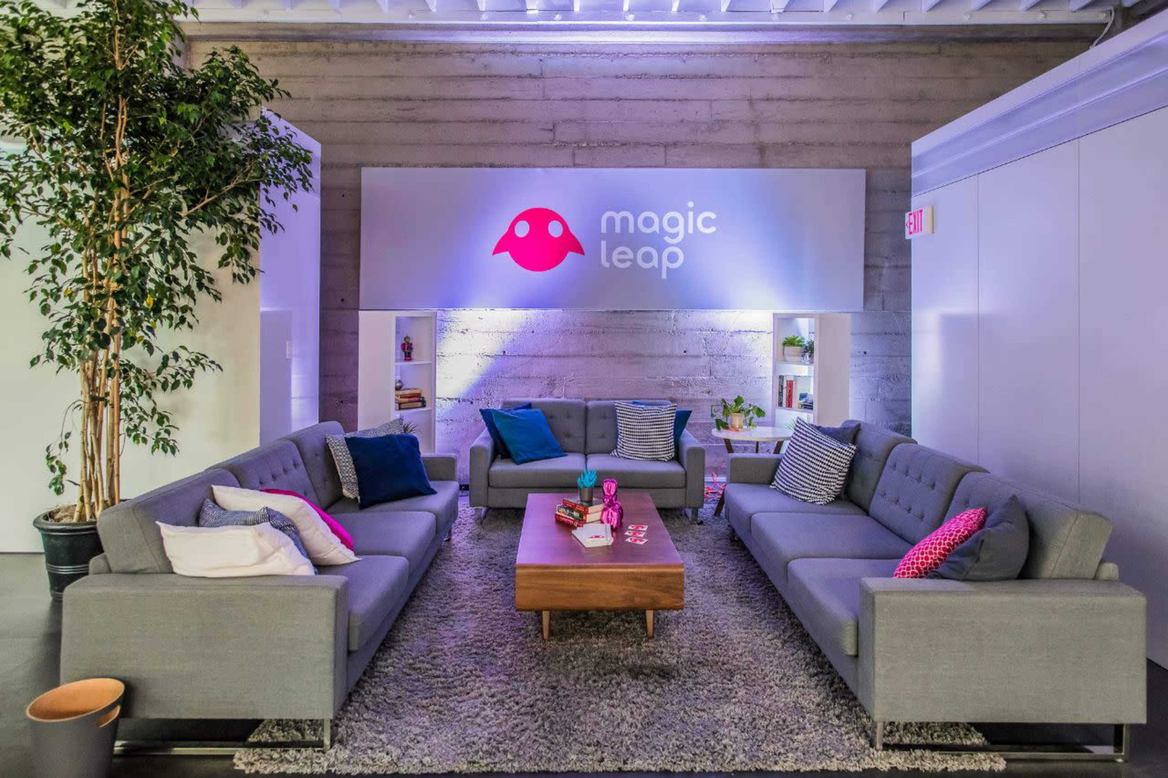 A modern lounge area featuring gray sofas, a wooden coffee table, and a decorative backdrop with the "Magic Leap" logo.