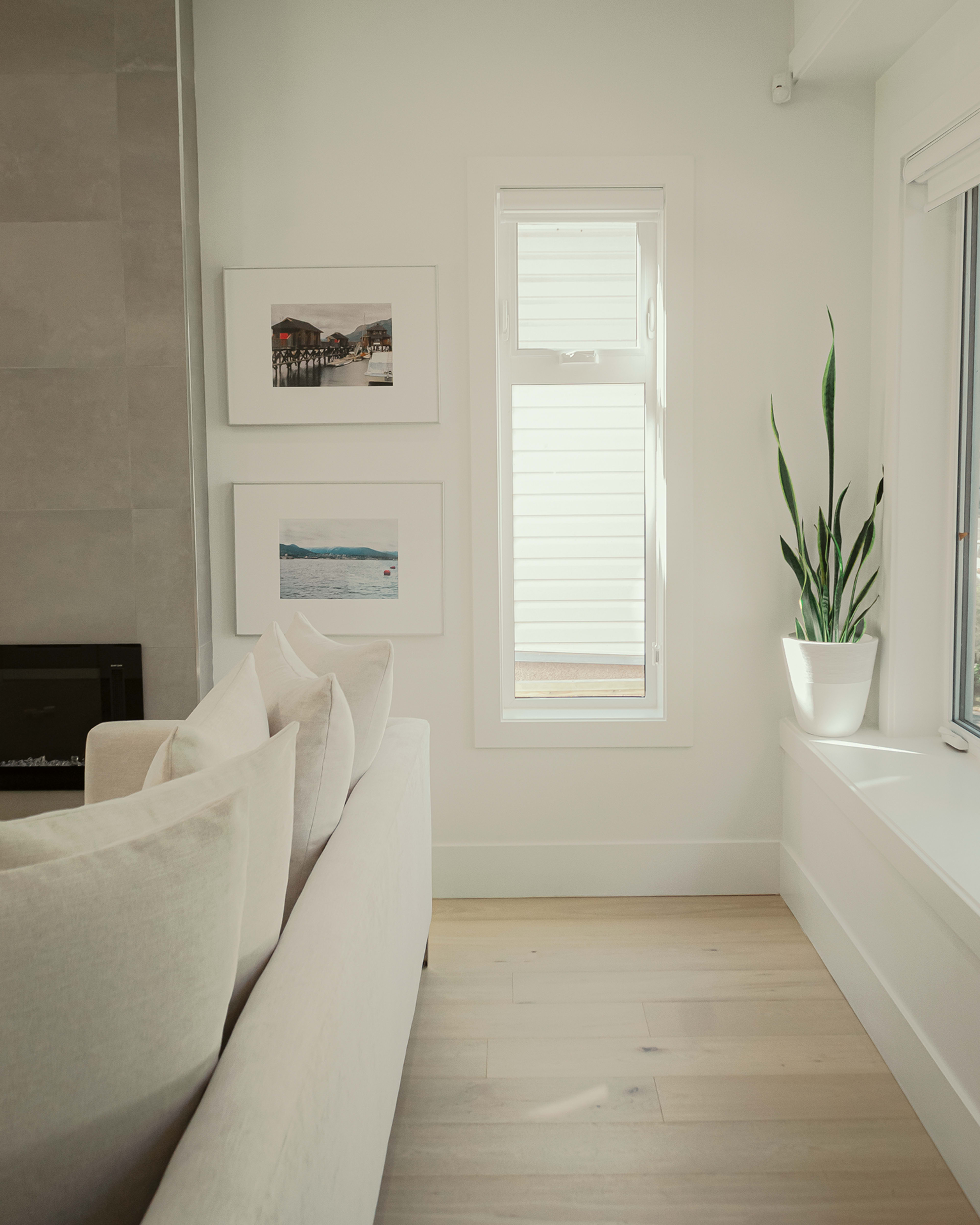 A minimalist living room with a white sofa, a potted plant on a windowsill, and framed pictures on the wall beside a window.