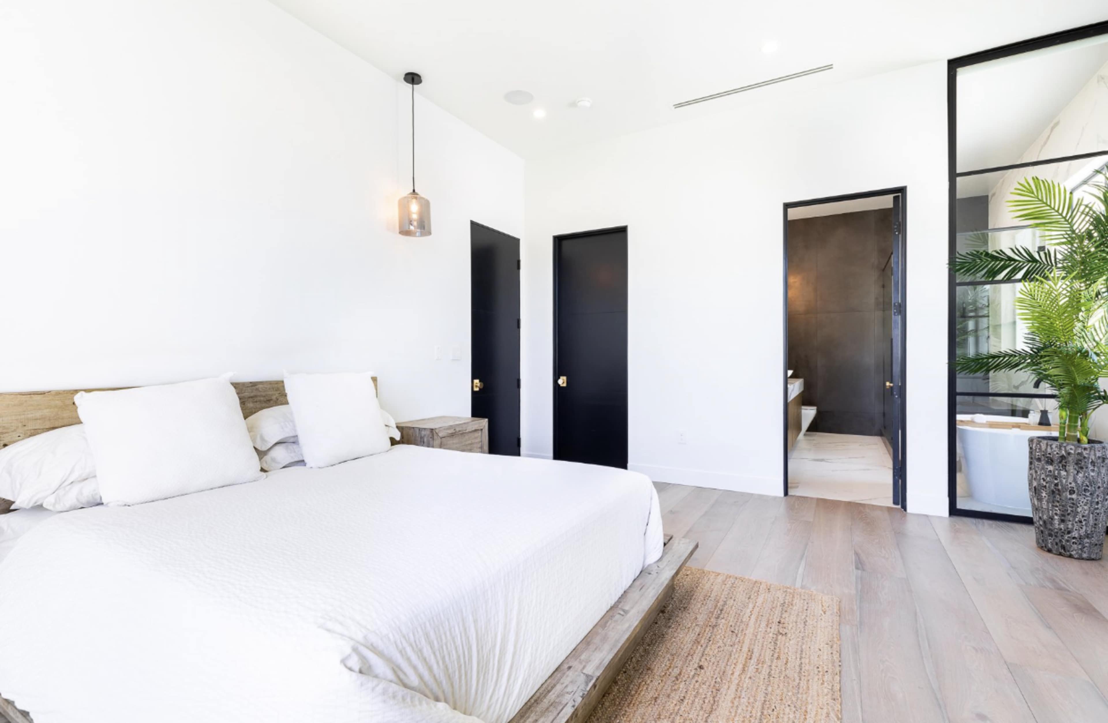A modern bedroom featuring a wooden bed frame, two white pillows, a jute rug, and a doorway leading to a bathroom with a black door and a glass wall.