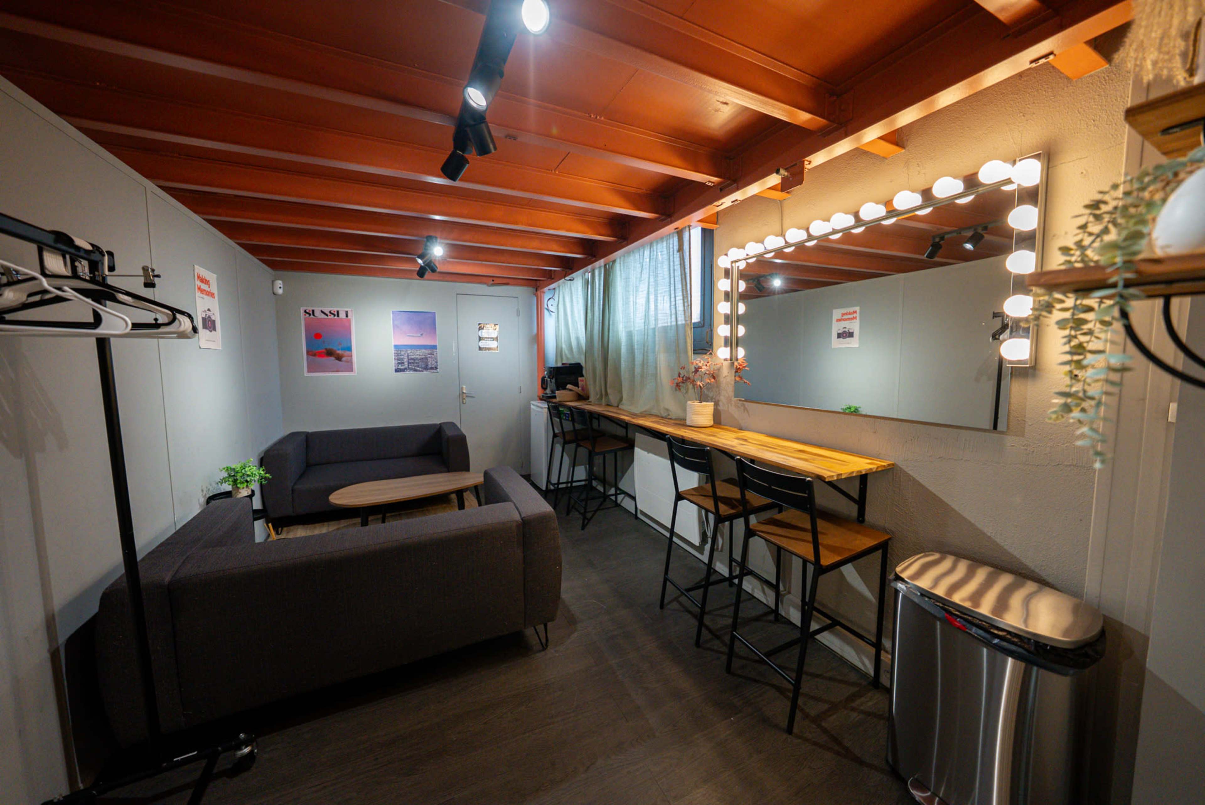The image shows a modern interior of a small lounge area featuring a gray sofa, a wooden table, a mirror with light bulbs, and a row of tall bar stools along a counter.