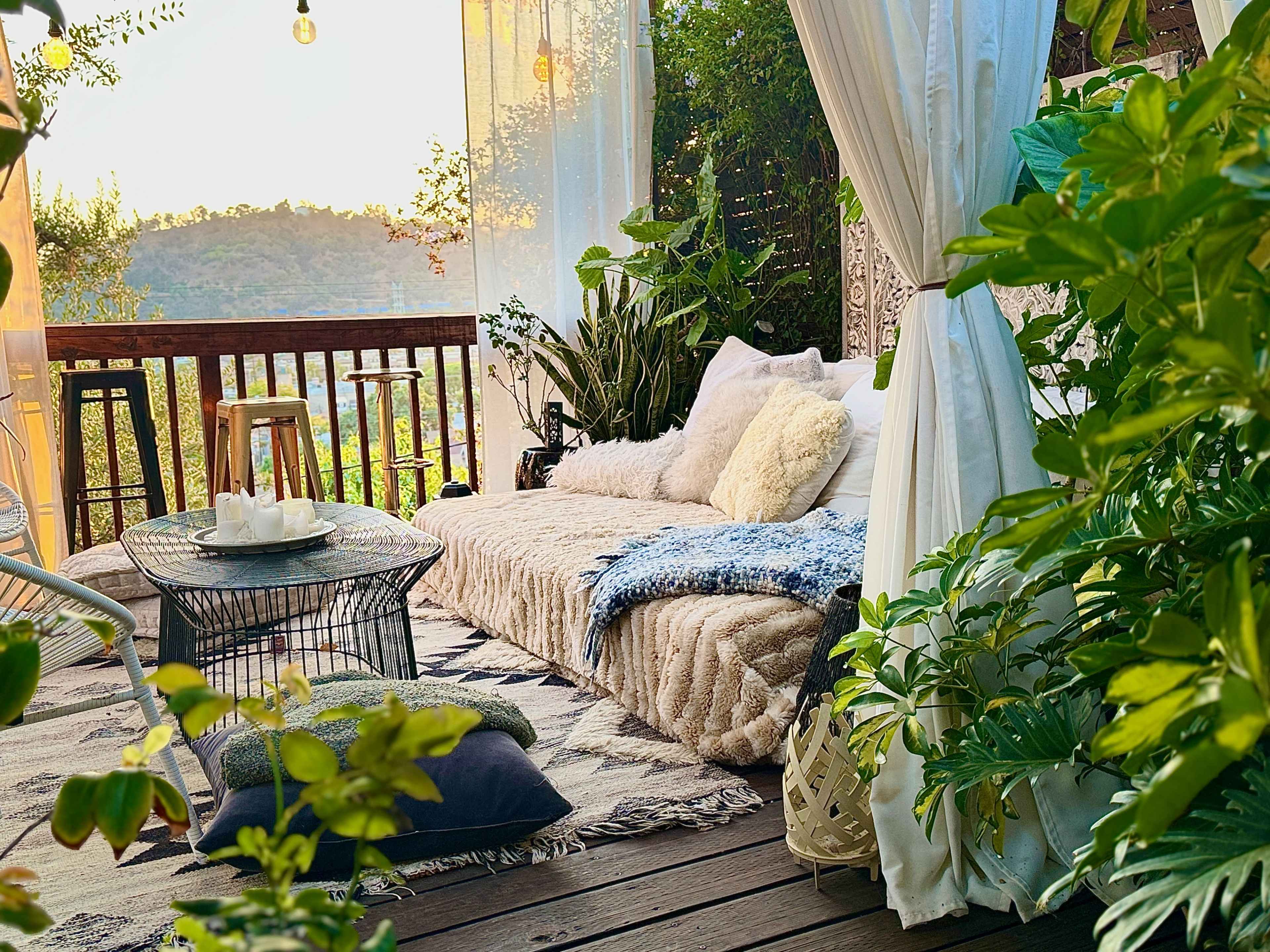 The image shows a cozy outdoor seating area adorned with plants, a plush couch, and a small table, set against a backdrop of distant hills and warm sunset light.