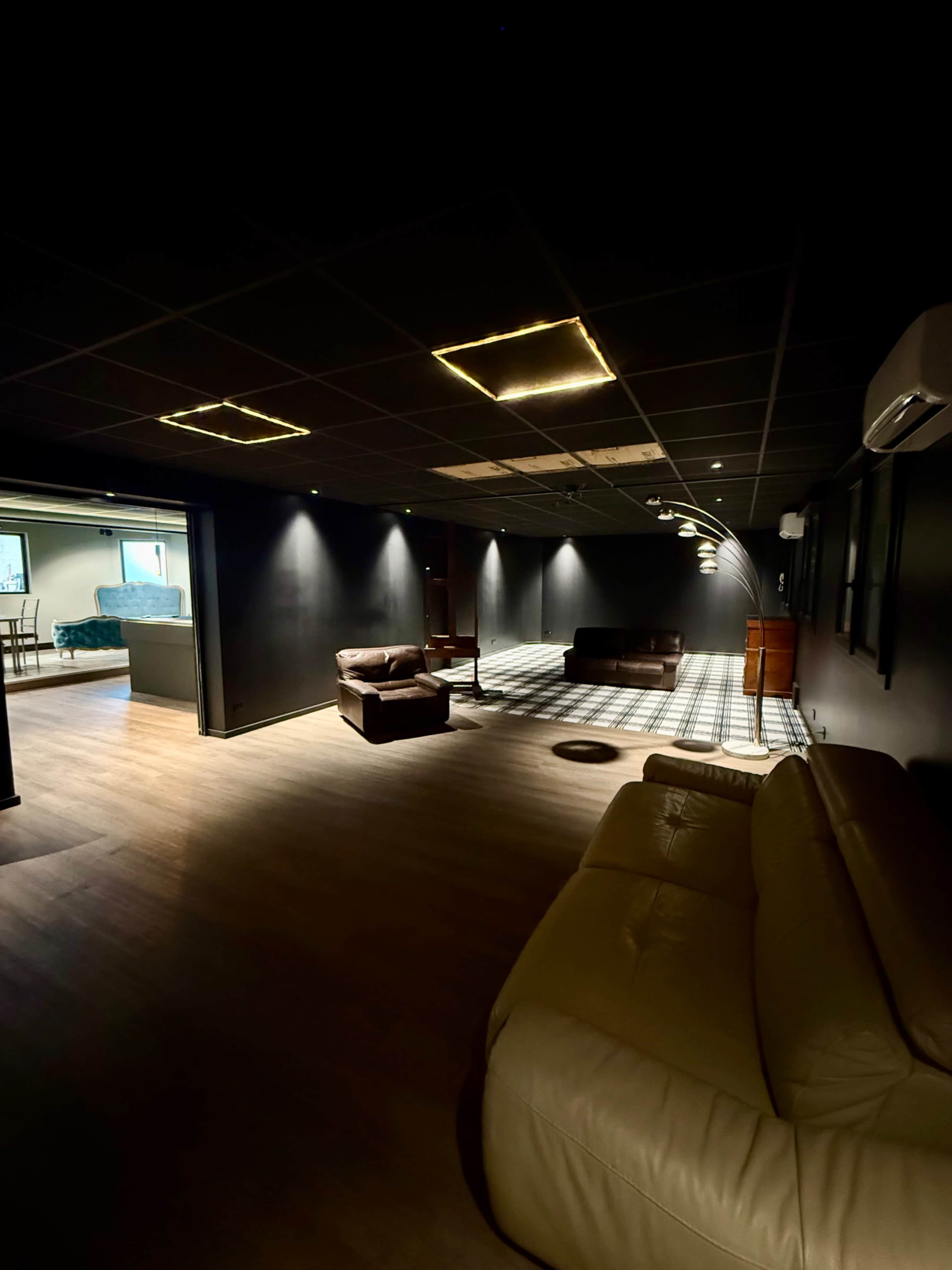 The image shows a dimly lit room with a dark ceiling, two couches, and a patterned floor leading to a brighter area filled with furniture.