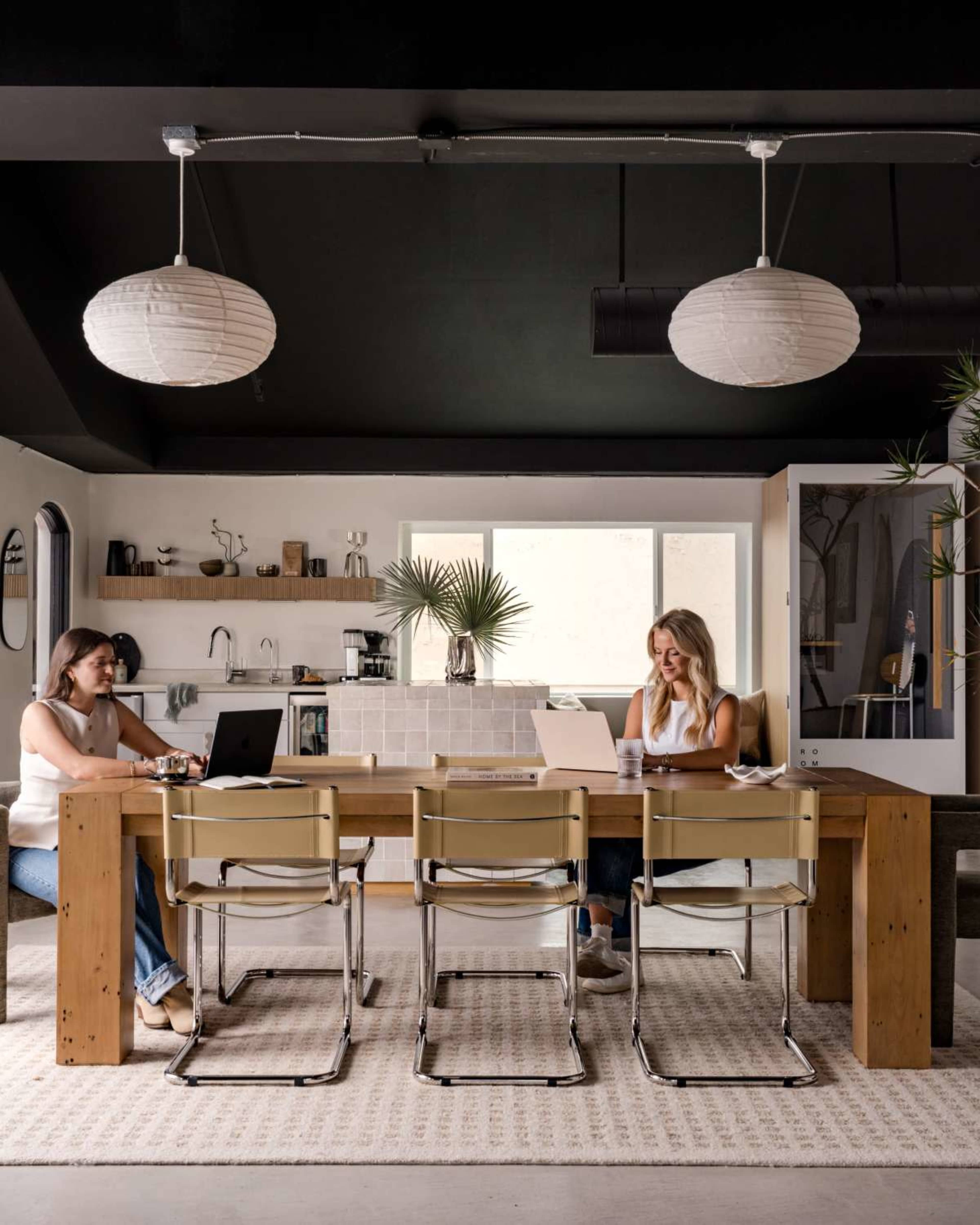 Two women sit at a large wooden table in a modern kitchen workspace, each using a laptop and one of them holding a mug.