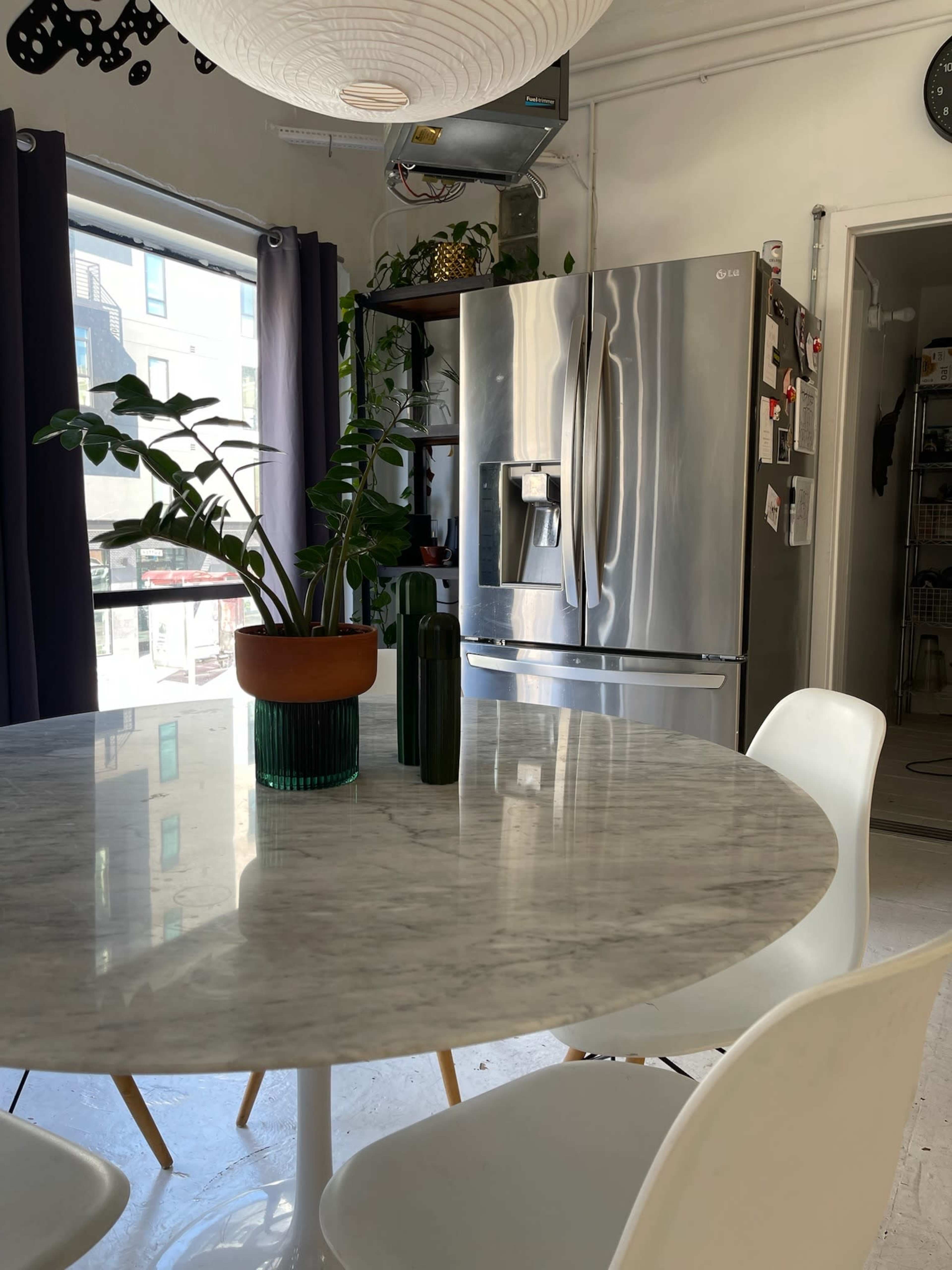 A round marble dining table with white chairs is positioned near a window, adjacent to a stainless steel refrigerator and a plant on the table.