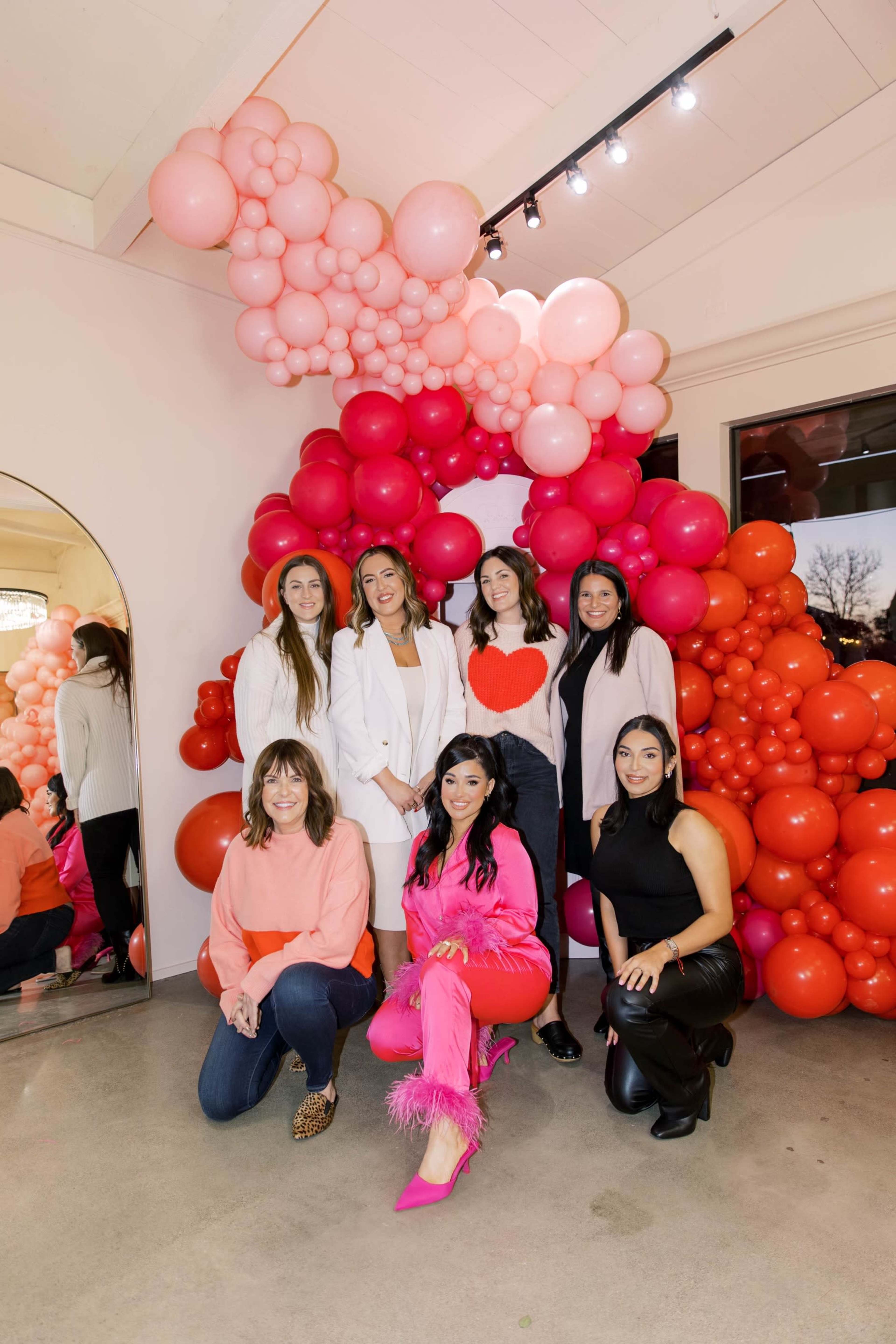 A group of six women poses for a photo in front of a colorful balloon display composed of various shades of pink and red.