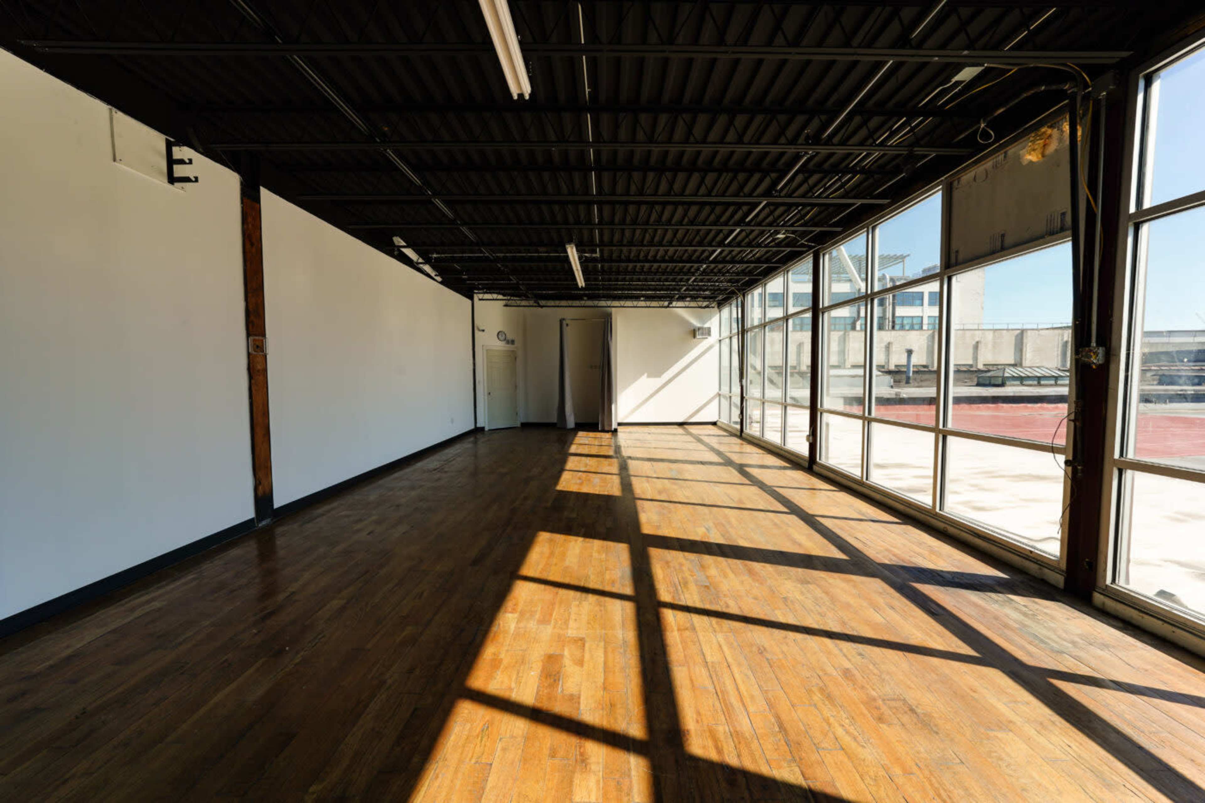 The image shows an empty room with hardwood floors and large windows allowing sunlight to fill the space.