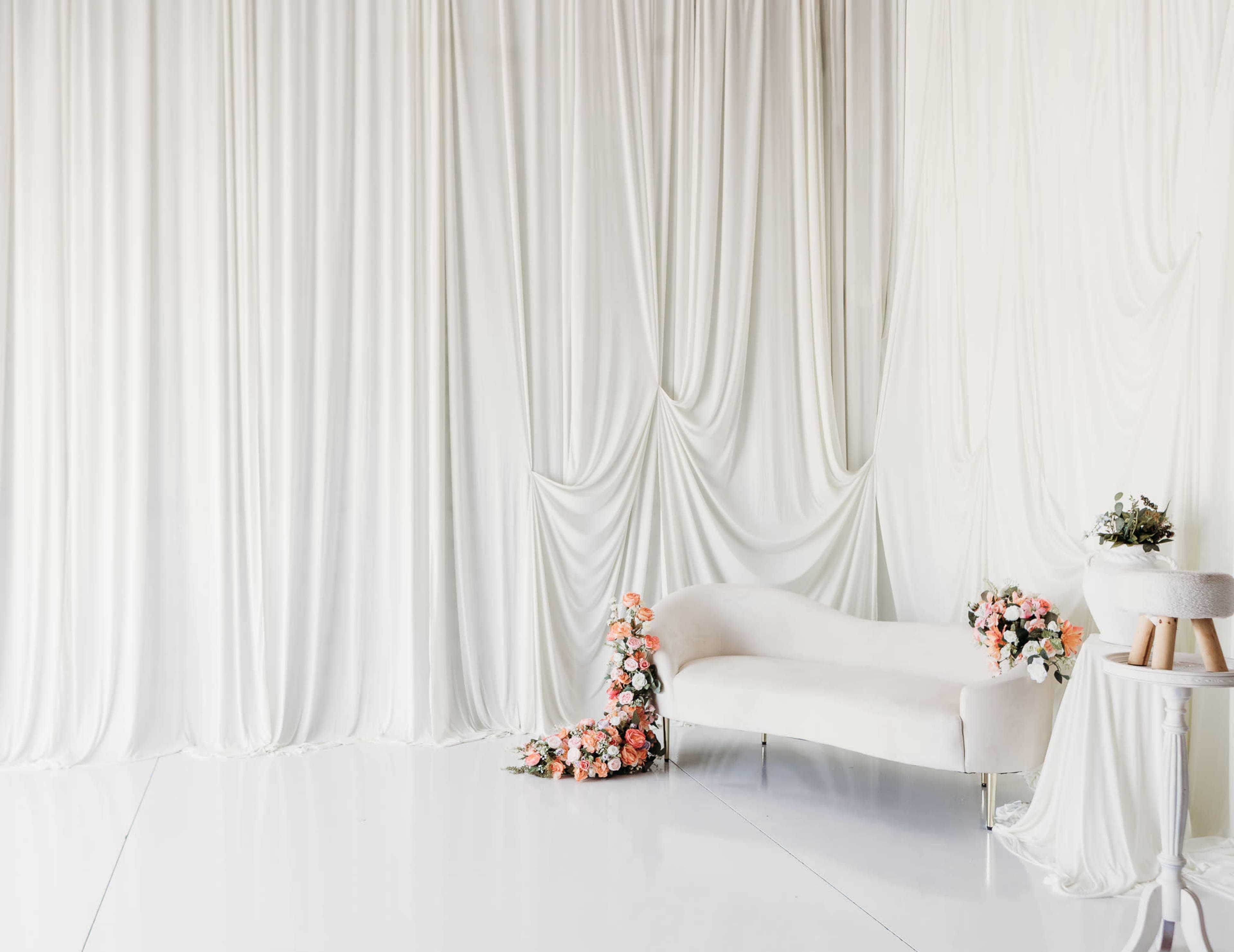 The image features a bright, minimalist interior with a white backdrop, a curved white sofa, and floral arrangements in soft pastel colors.