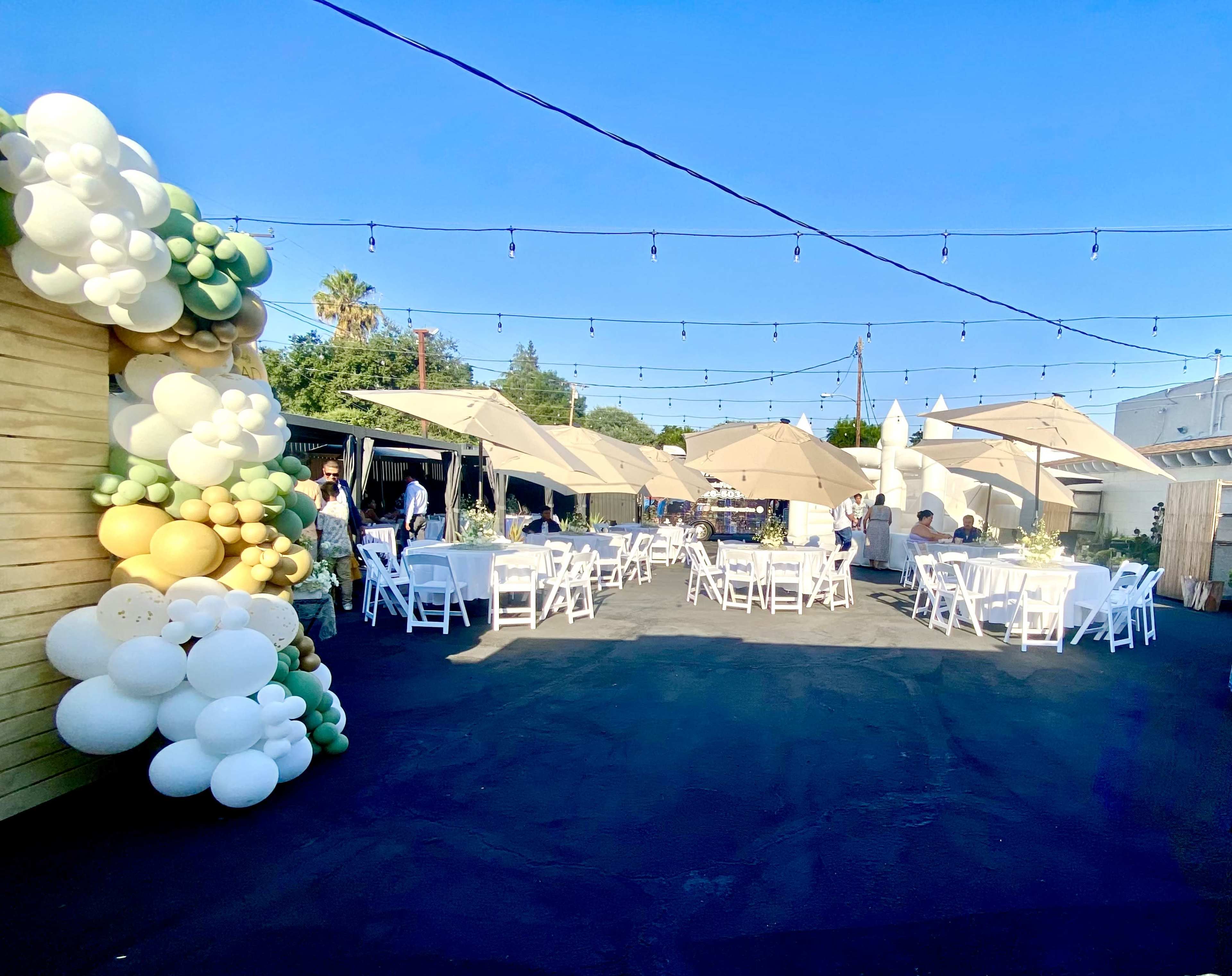The image shows a decorated outdoor event space with tables set under large umbrellas and a colorful balloon arch on one side.