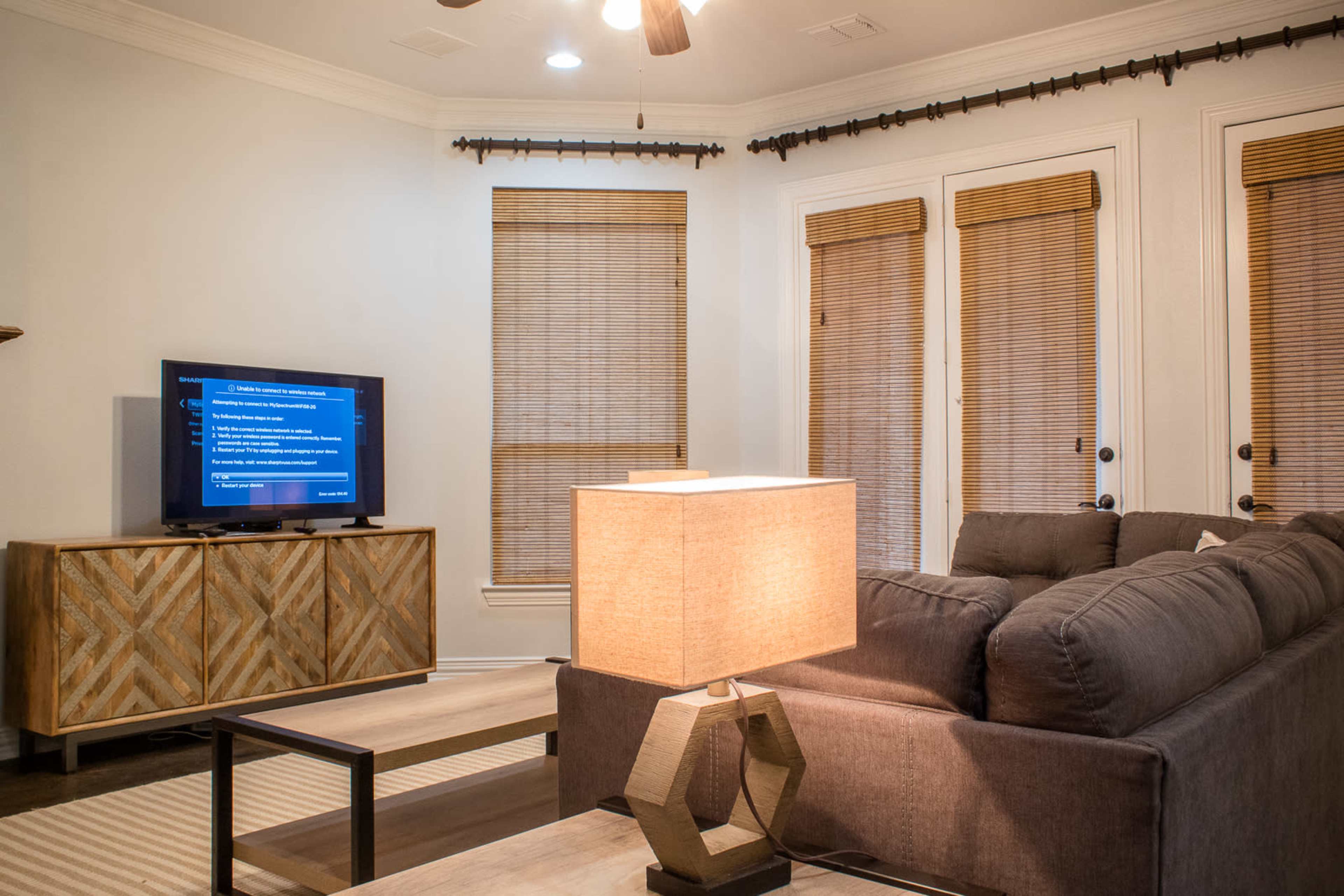 A living room features a sofa, coffee table, and a television displaying content, with natural light filtering through window blinds.