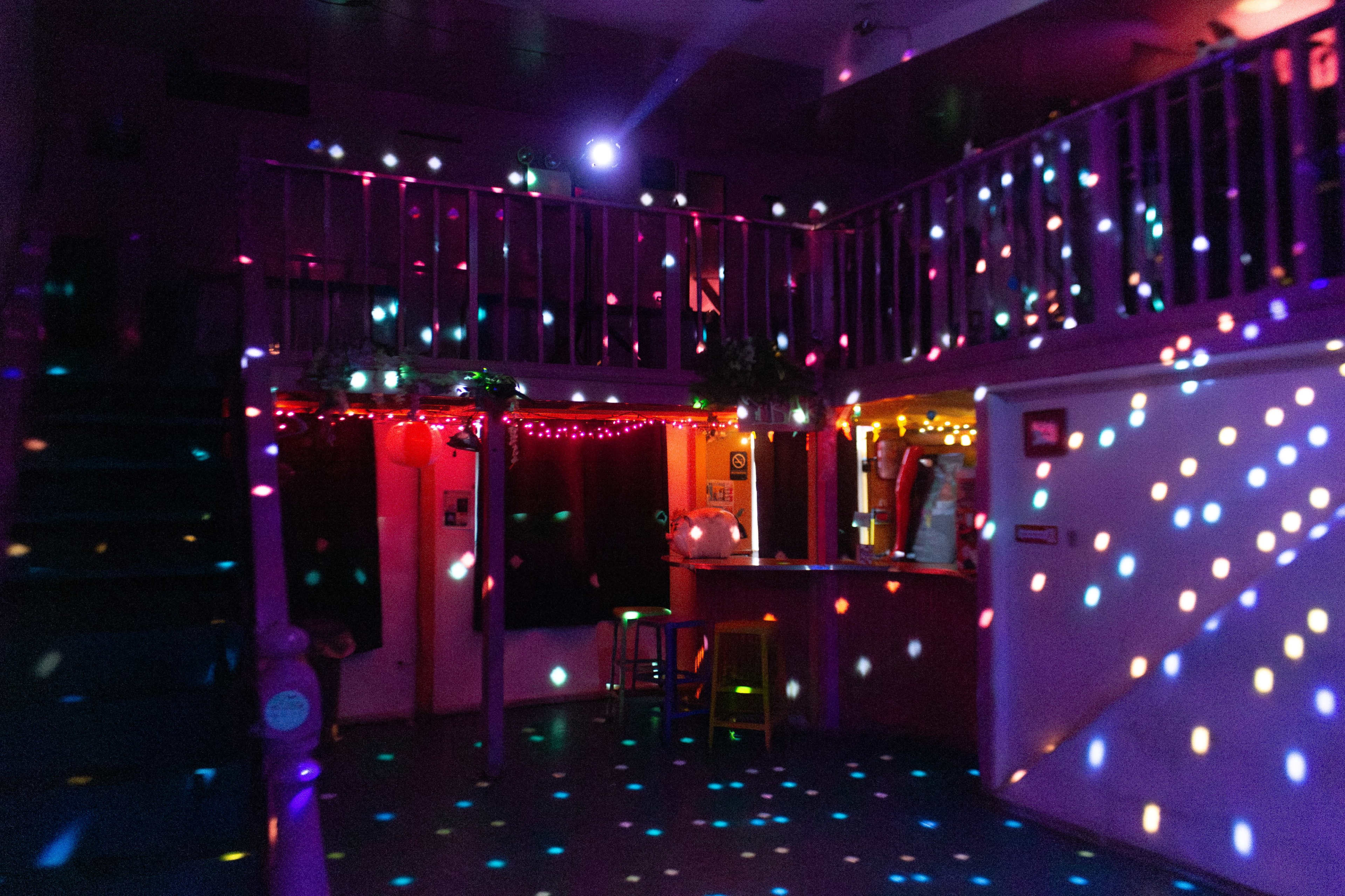 The image shows a dimly lit room with colorful lights casting circular patterns on the floor and walls, a bar area, and a staircase in the background.