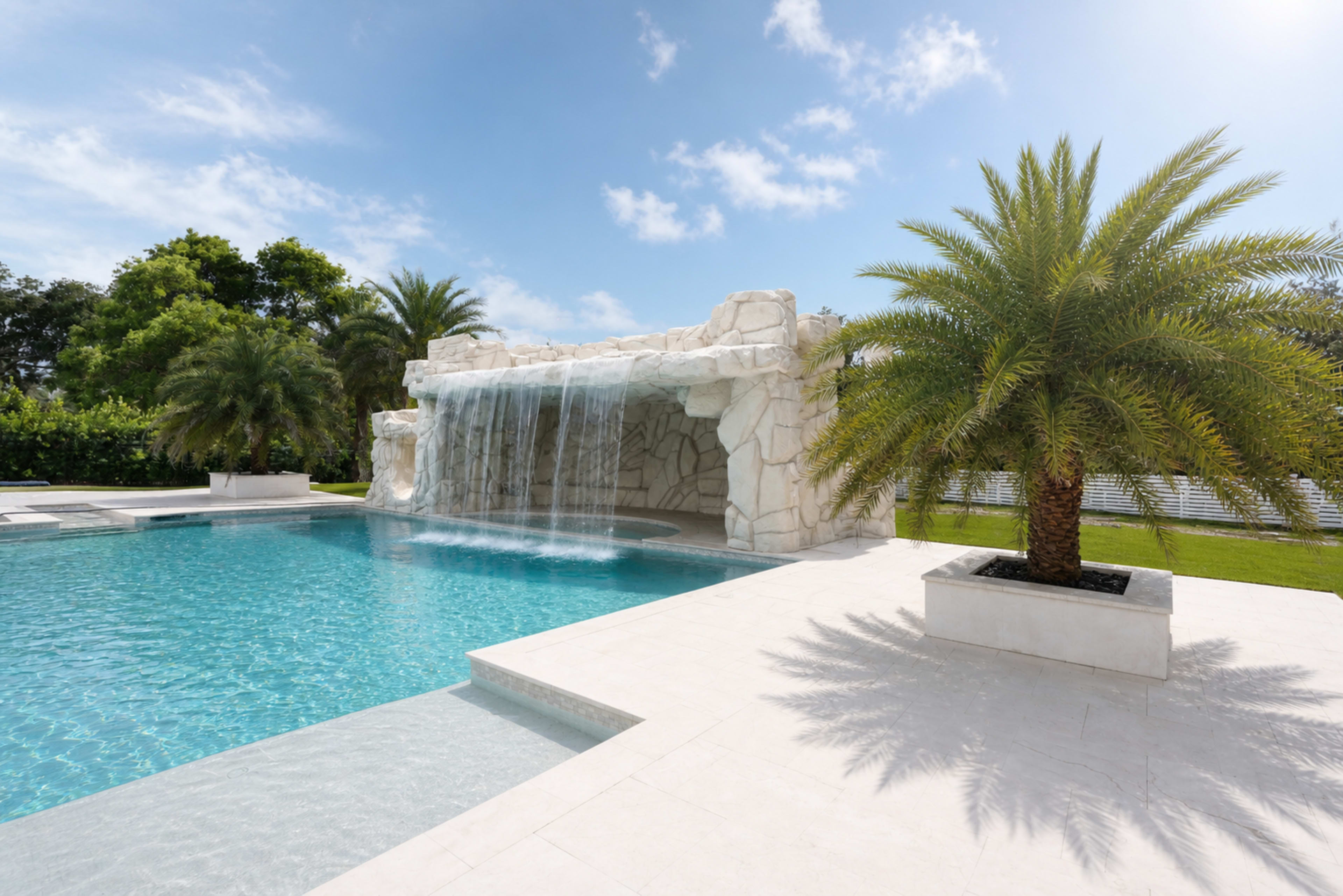 The image shows a luxurious swimming pool with a waterfall feature, surrounded by palm trees and a clear blue sky.