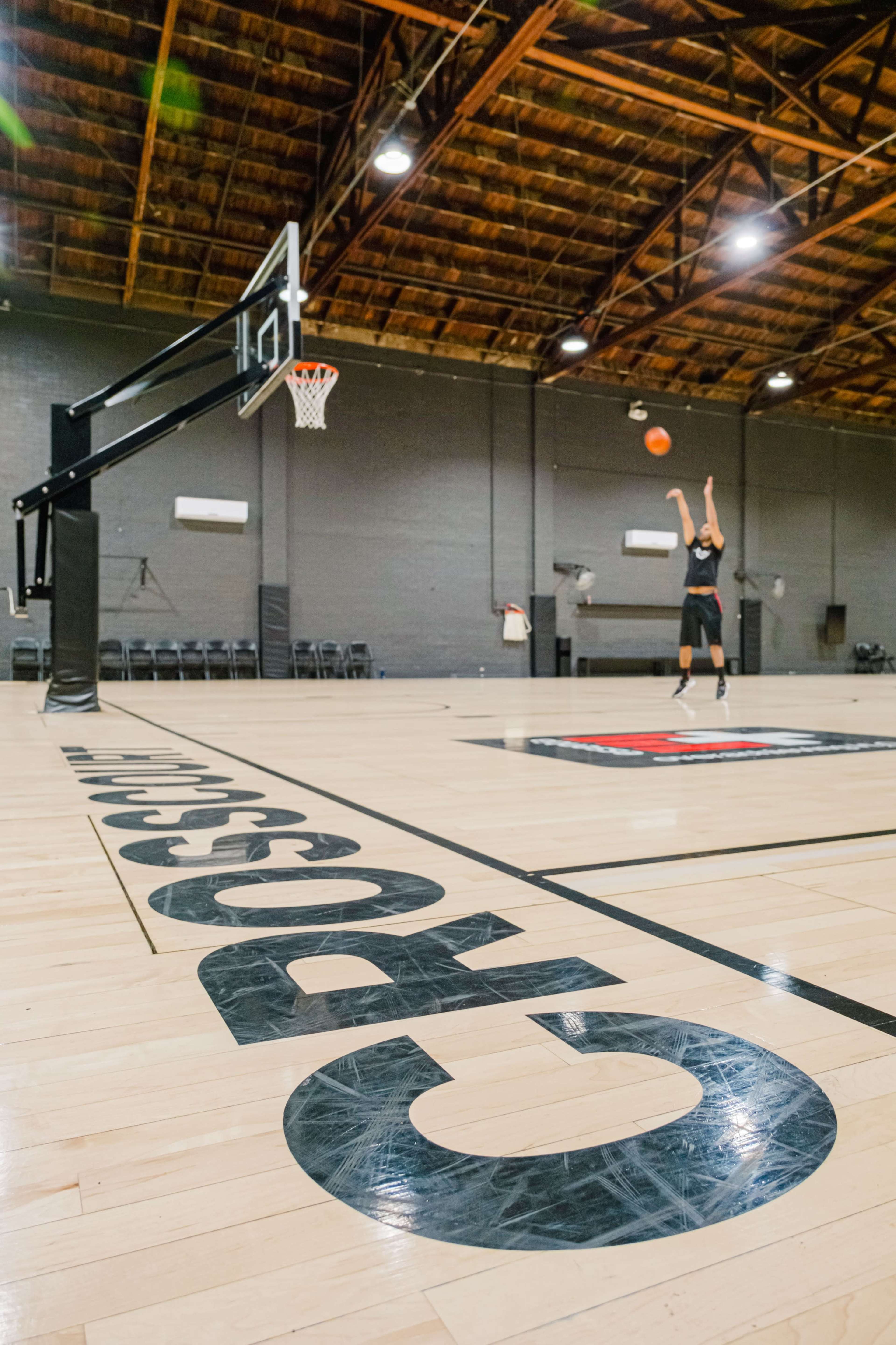 A player takes a jump shot on a basketball court featuring a prominent logo and empty seating in the background.