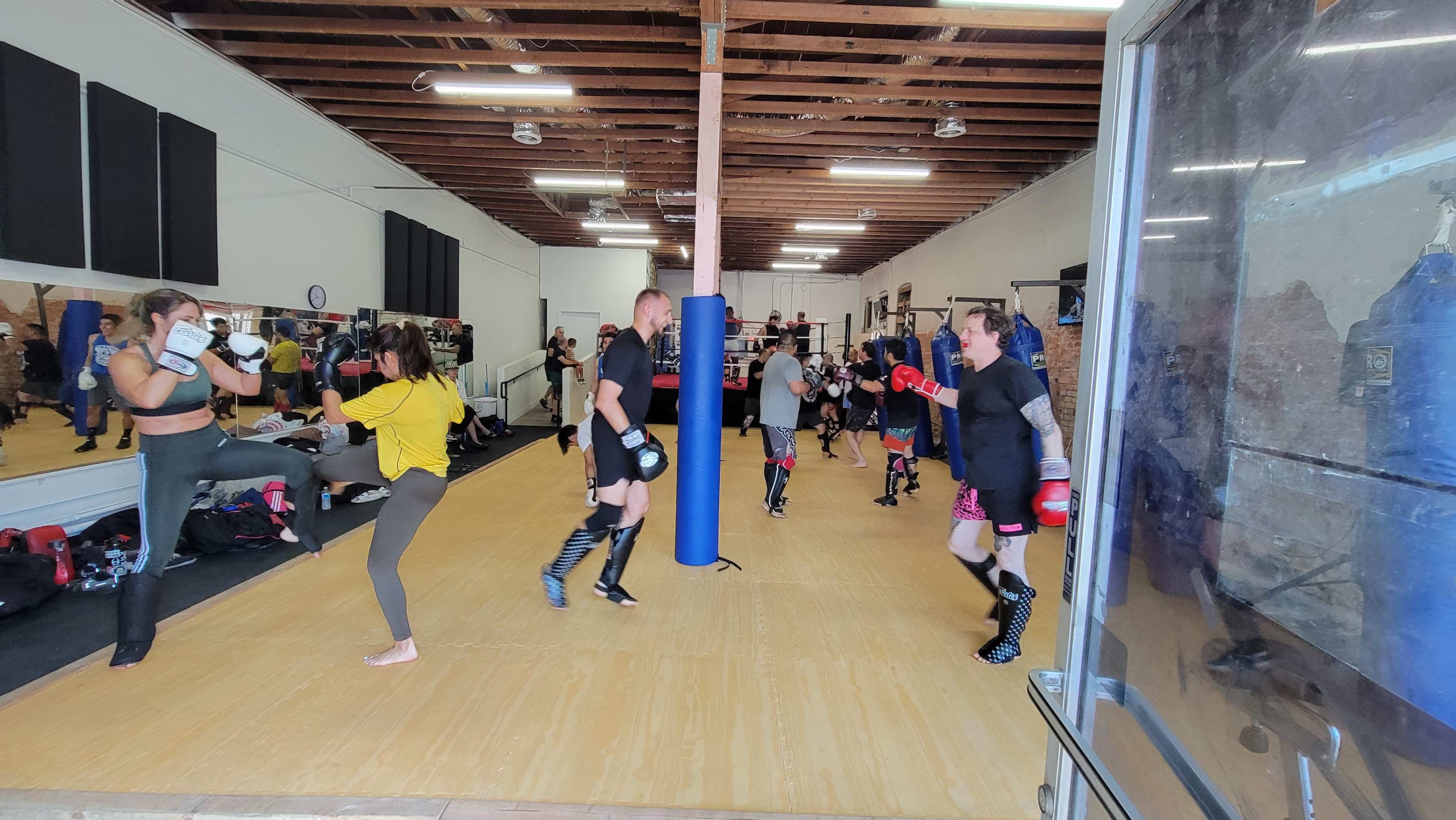 A group of individuals is training in a martial arts gym, practicing boxing and kickboxing techniques in a spacious room with equipment and mirrors.