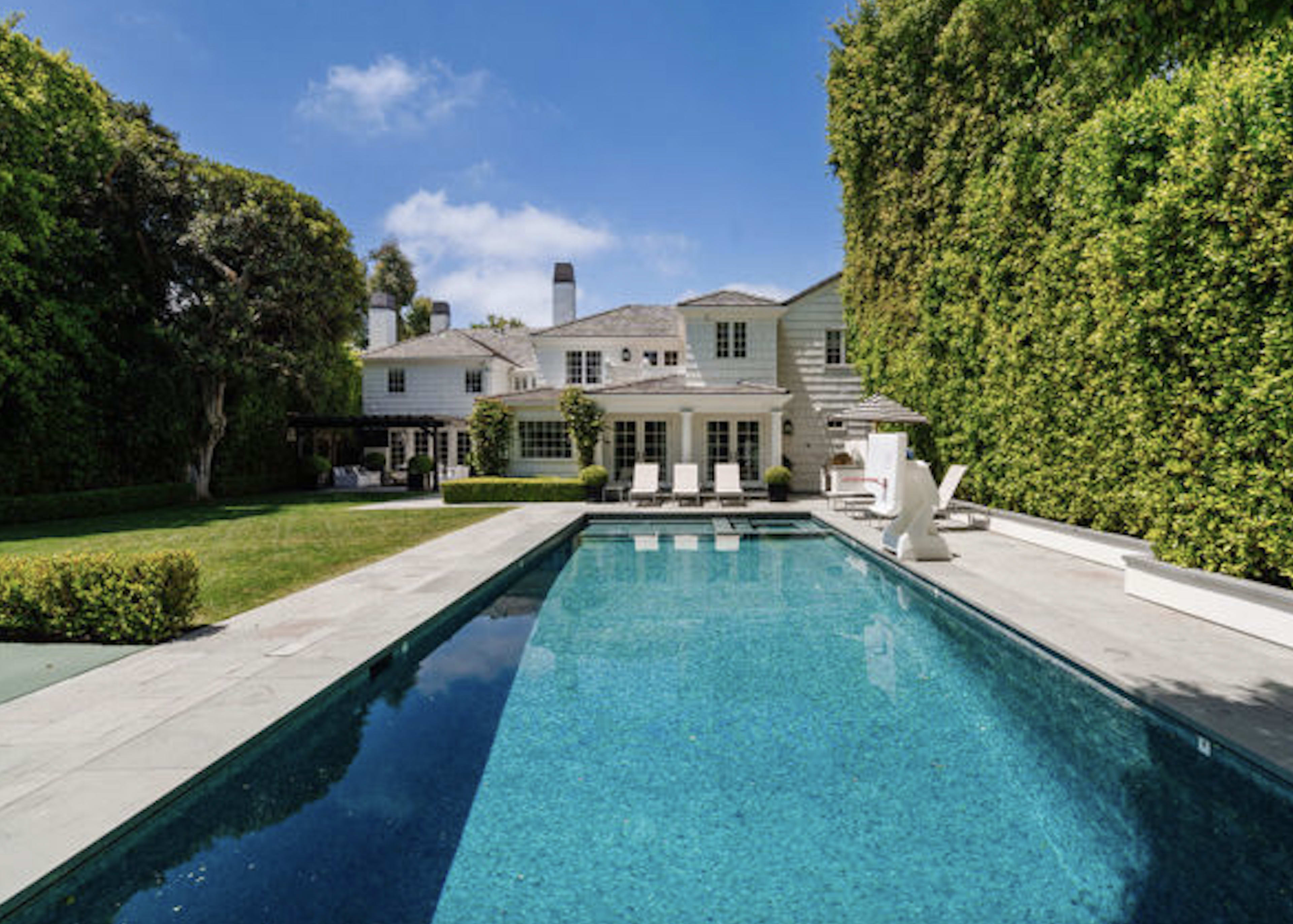 The image shows a spacious backyard with a swimming pool, framed by tall hedges and a large house in the background.