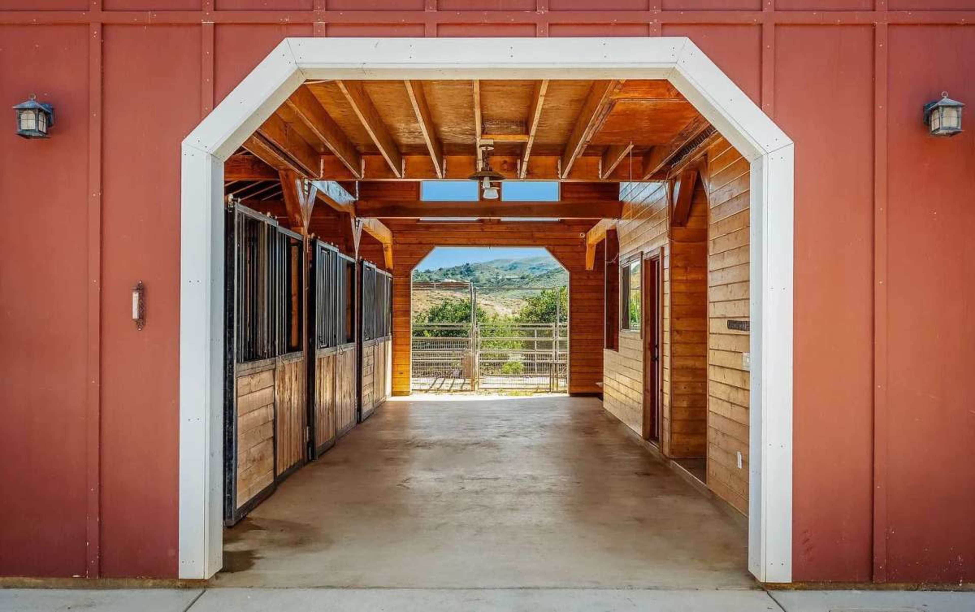 The image shows a rectangular barn entrance that opens to a spacious interior with wooden walls, exposing a view of rolling hills in the background.