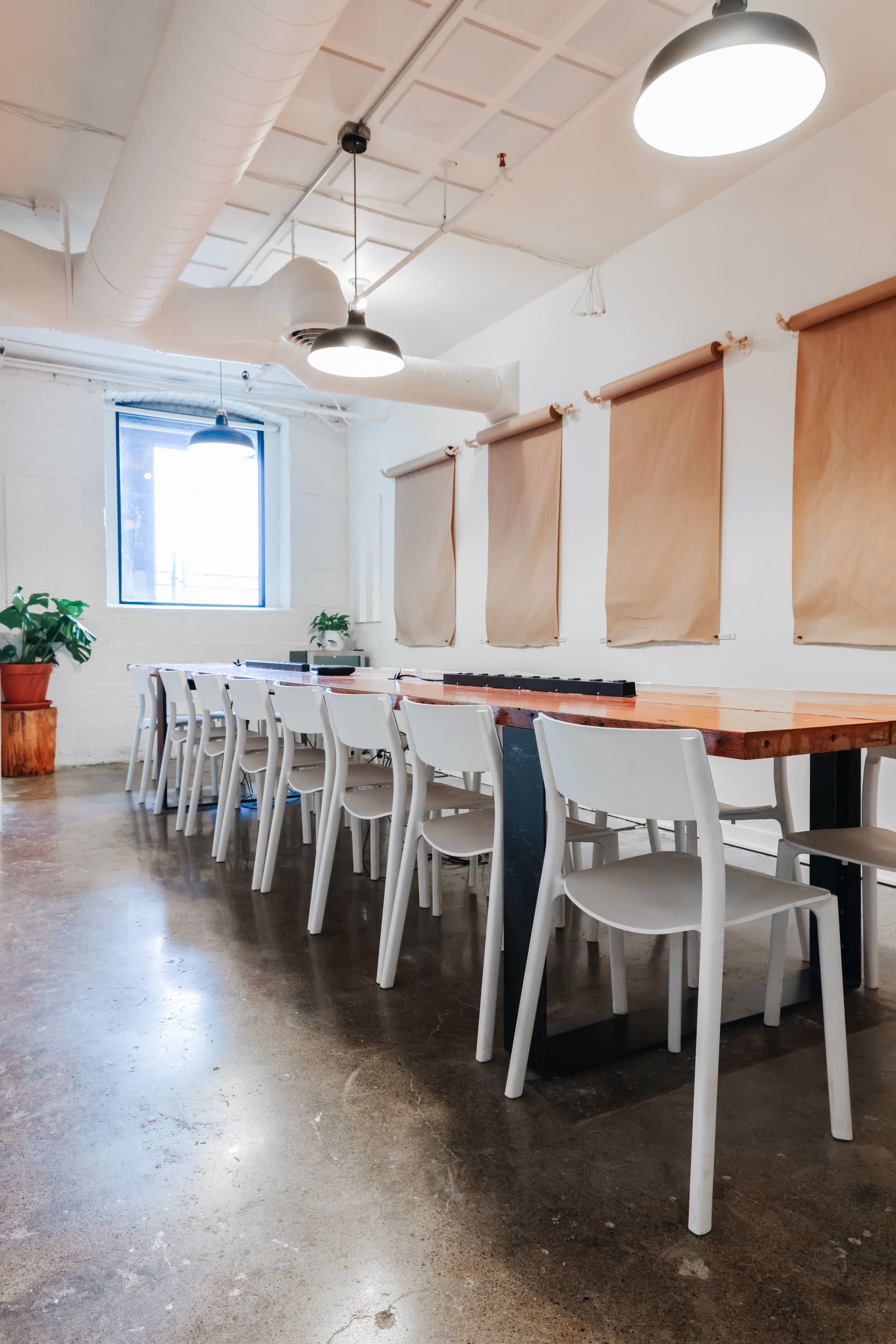 A long wooden table with white chairs is arranged in a well-lit room with large windows and hanging light fixtures.