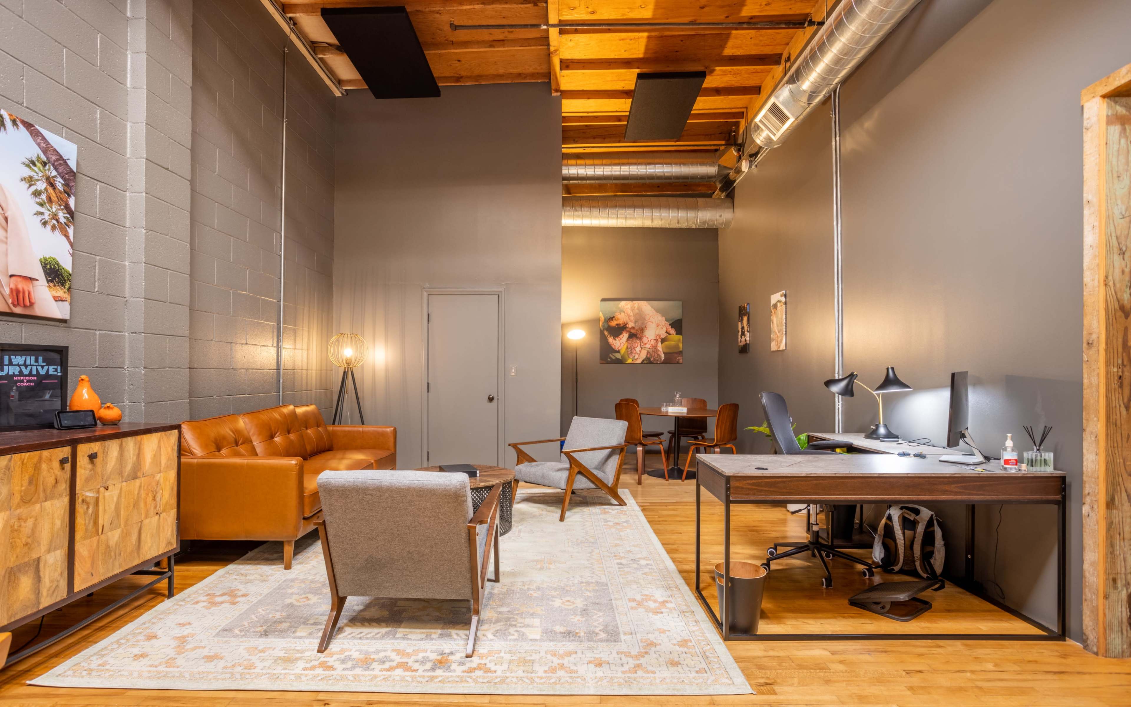 A modern office space featuring a brown leather sofa, a wooden desk with a lamp, and a decorative rug on a hardwood floor.
