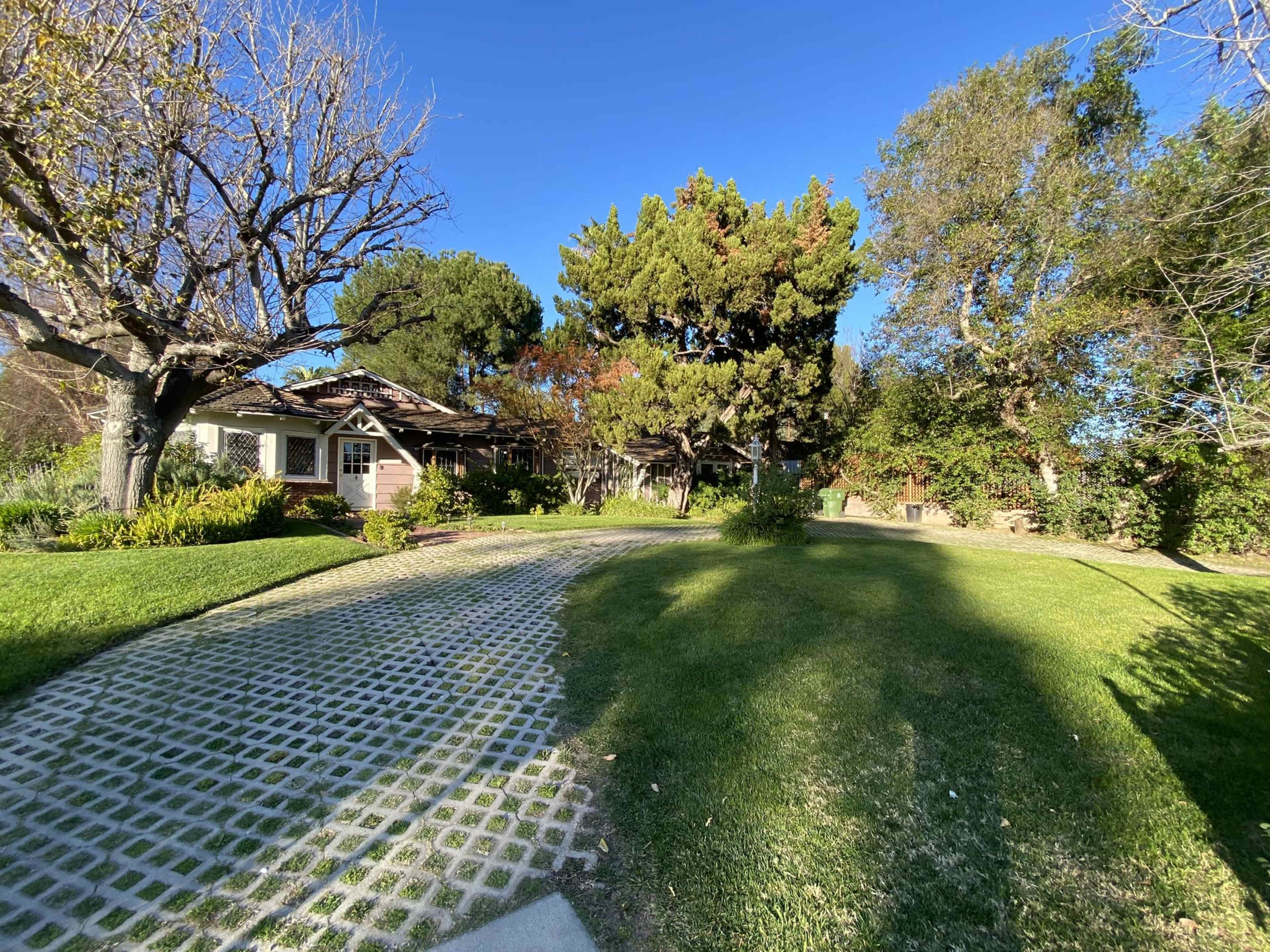 A landscaped garden with a cobblestone pathway, surrounded by various trees and a house in the background.